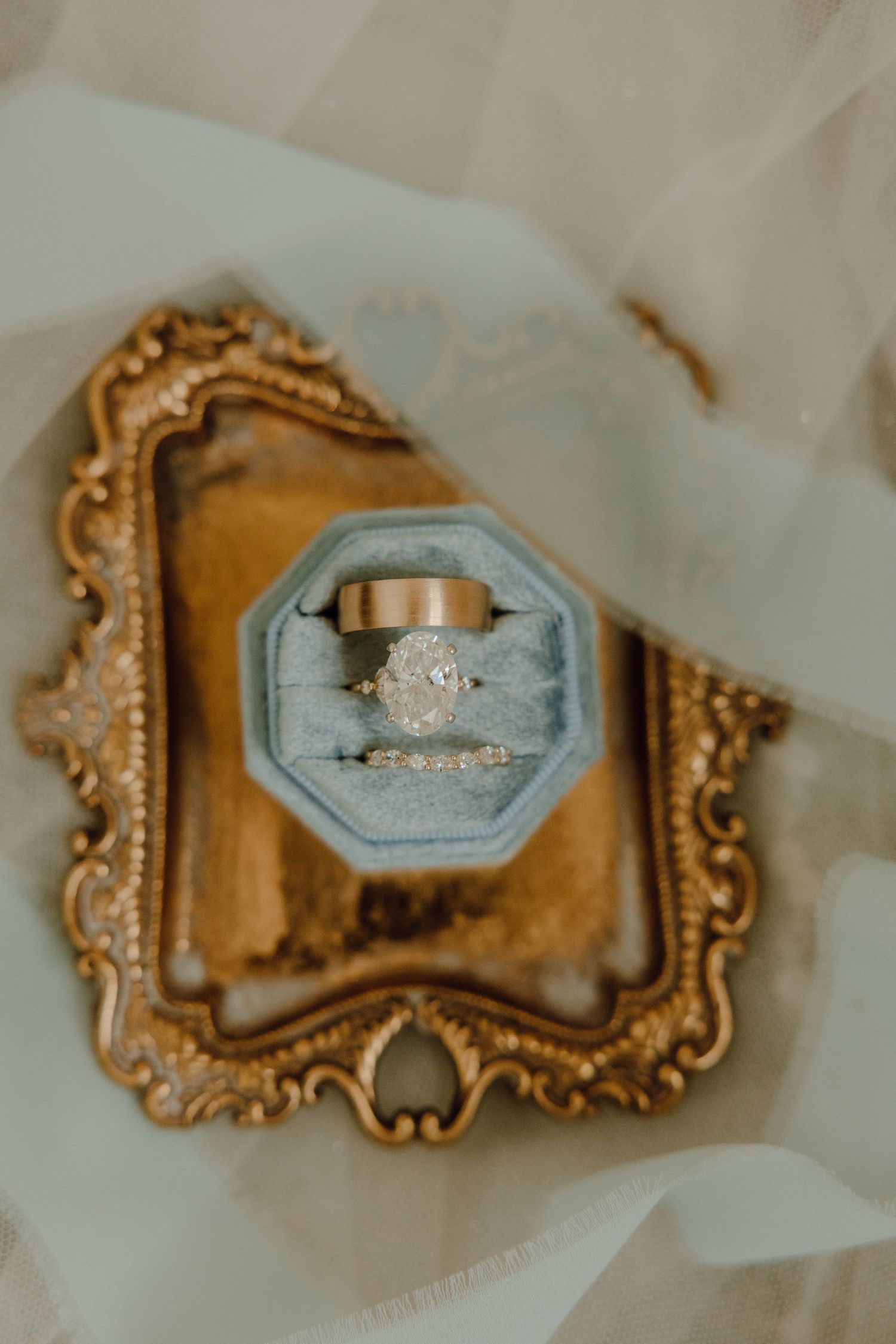 Elegant oval diamond engagement ring with wedding bands displayed in a gray velvet ring box on a gold ornate tray.