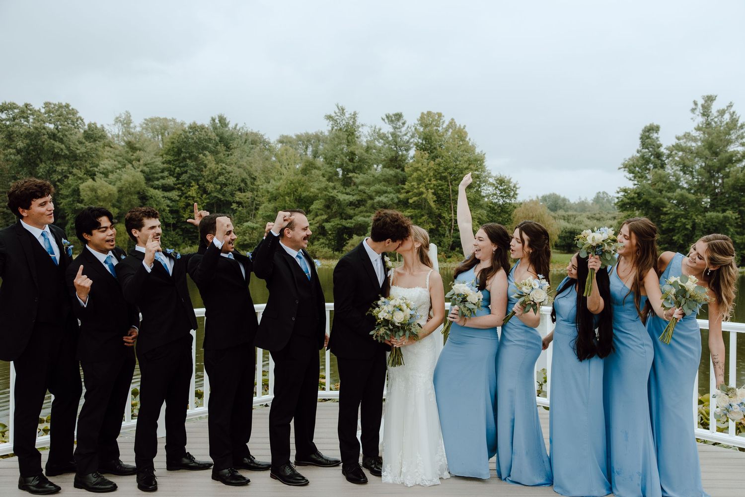 Wedding party in black suits and blue bridesmaid dresses celebrating on an outdoor deck surrounded by lush greenery.