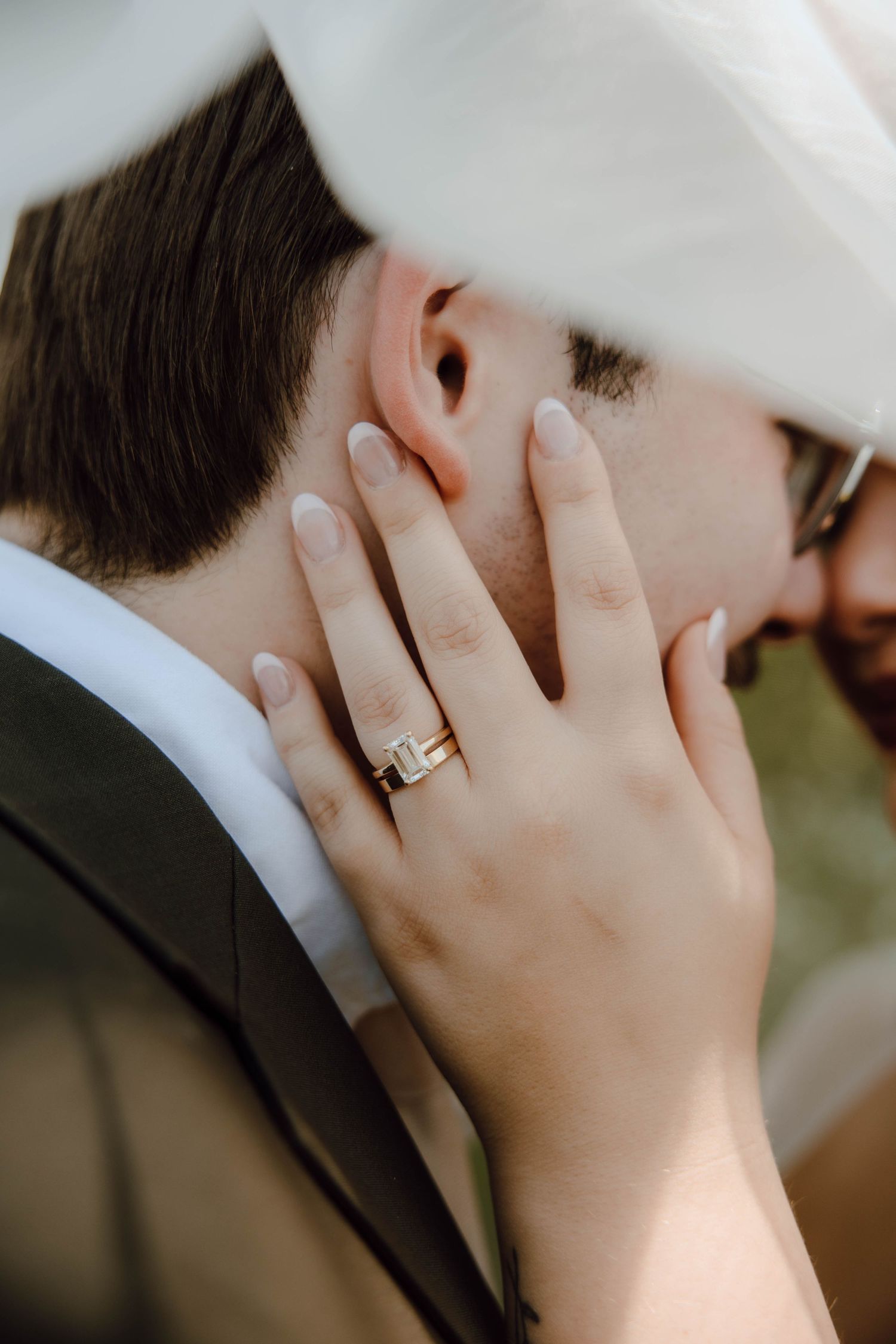 Bride's hand with gold emerald-cut wedding ring gently cupping groom's face under white veil.