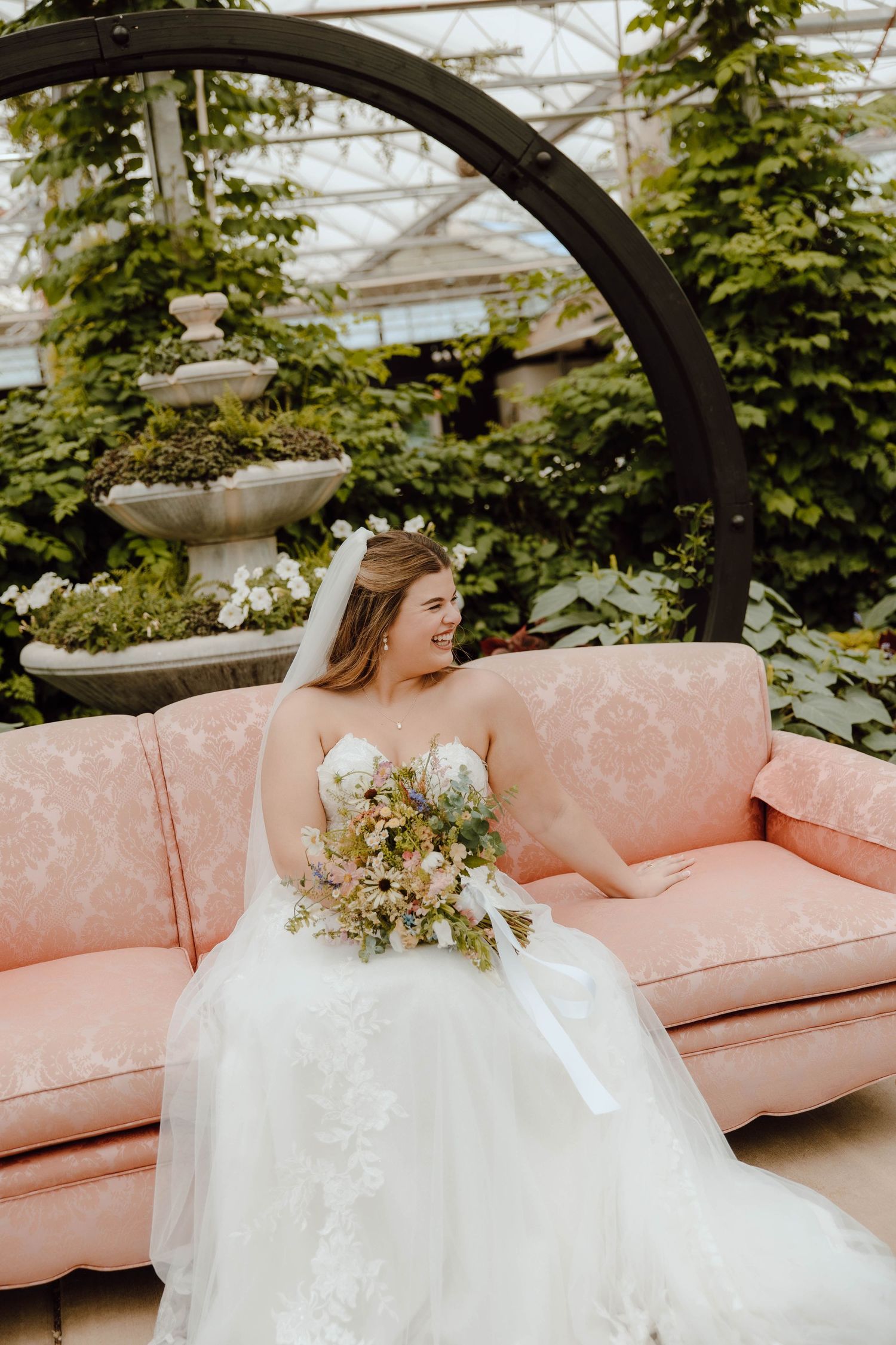 Bride in lace ballgown sits on pink damask sofa holding floral bouquet in greenhouse wedding venue.