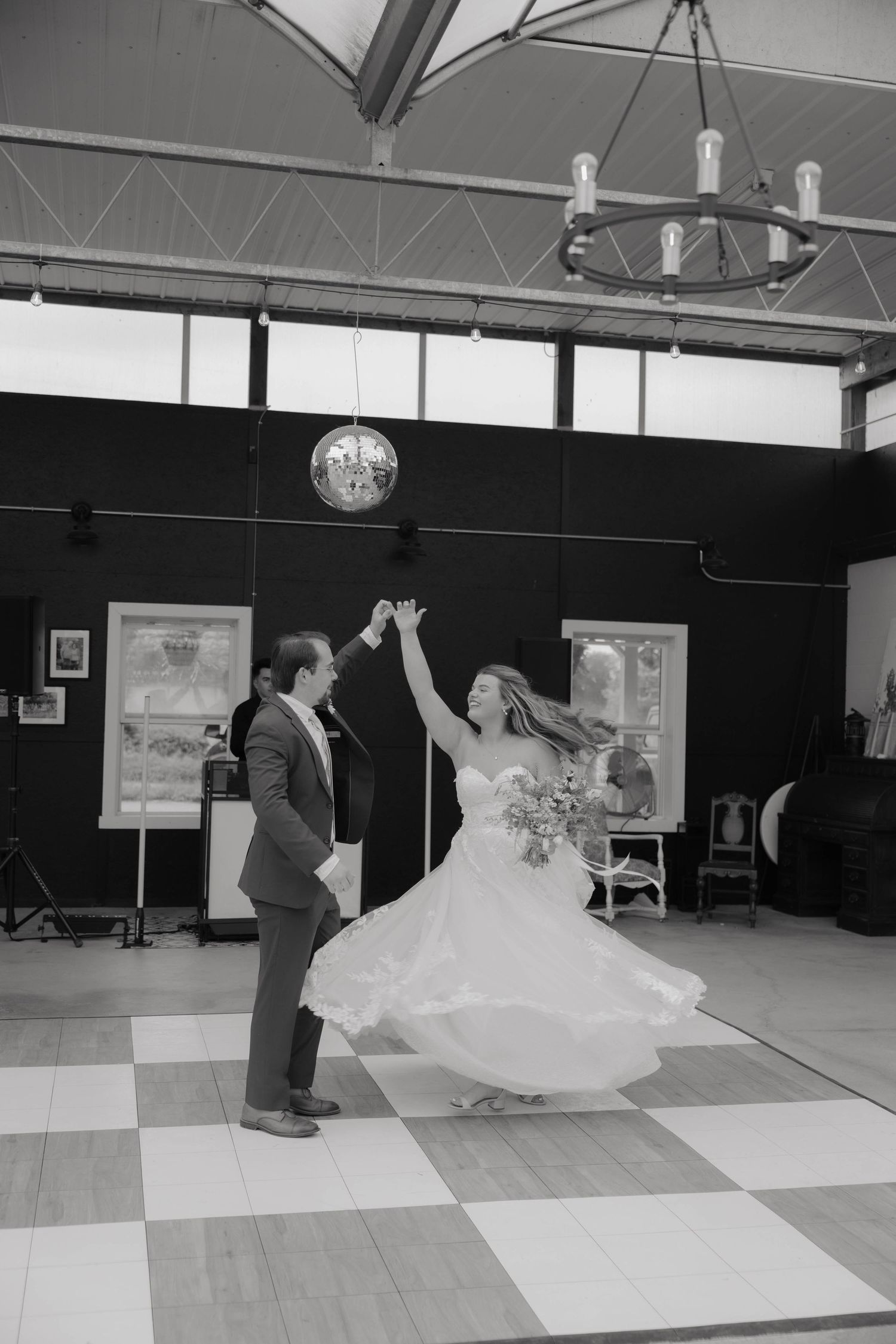 Bride and groom twirling together under disco ball on checkered dance floor in black and white.