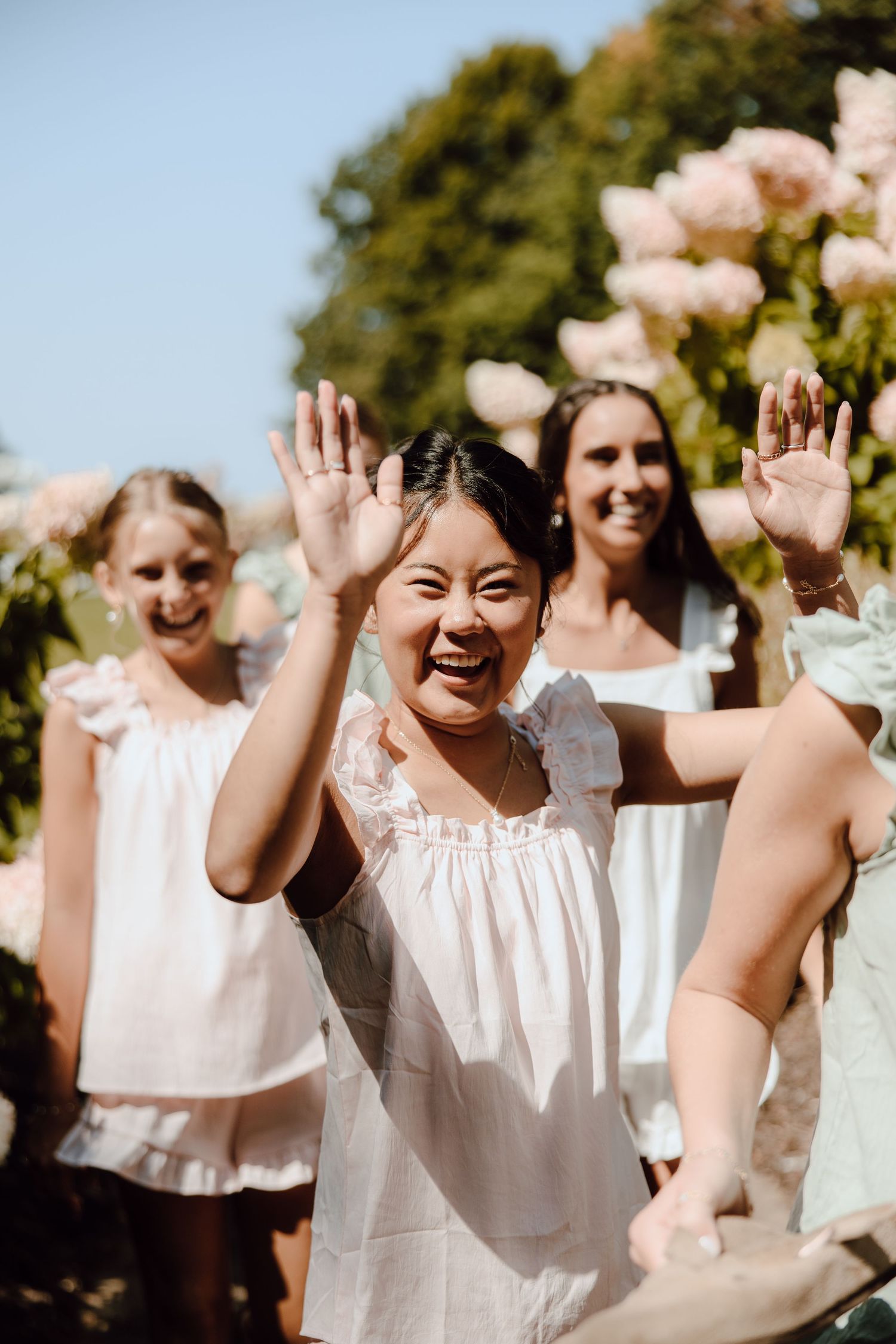 Joyful woman in white dress waves both hands outdoors near pink flowers with smiling friends behind her.