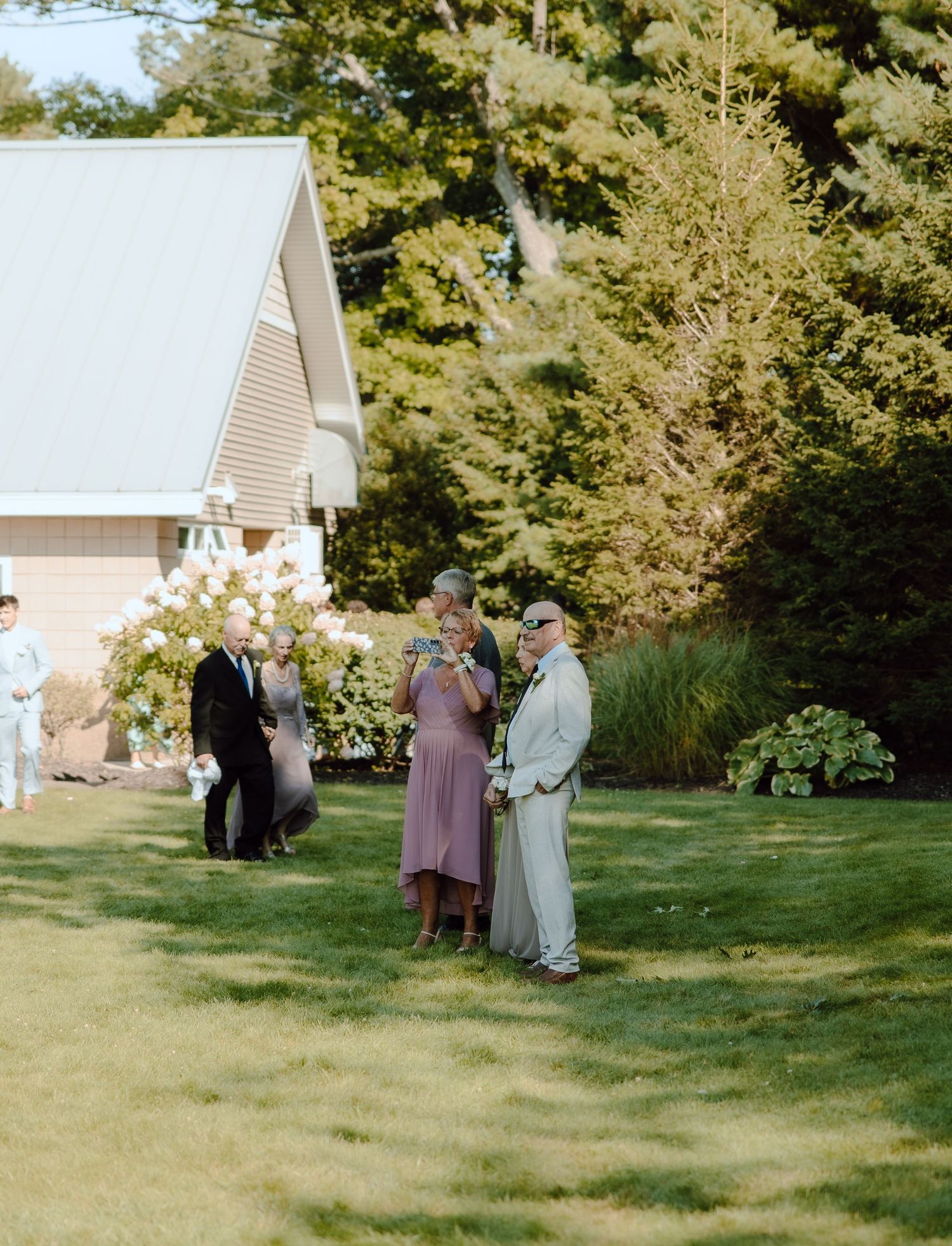 Outdoor wedding ceremony on green lawn with guests in formal attire near a white building surrounded by lush trees.