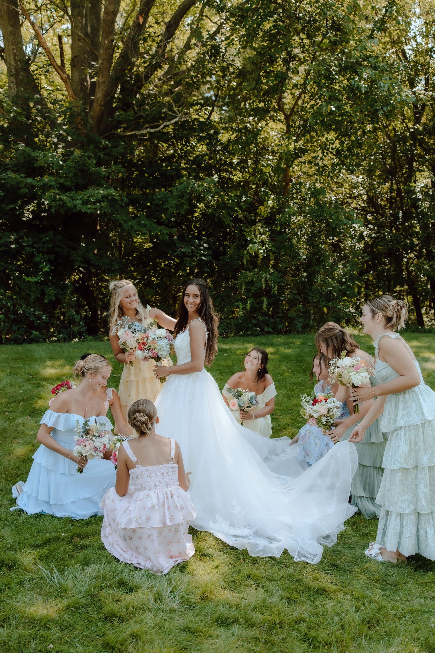 Bride surrounded by bridesmaids in mismatched dresses holding colorful bouquets on a green lawn.