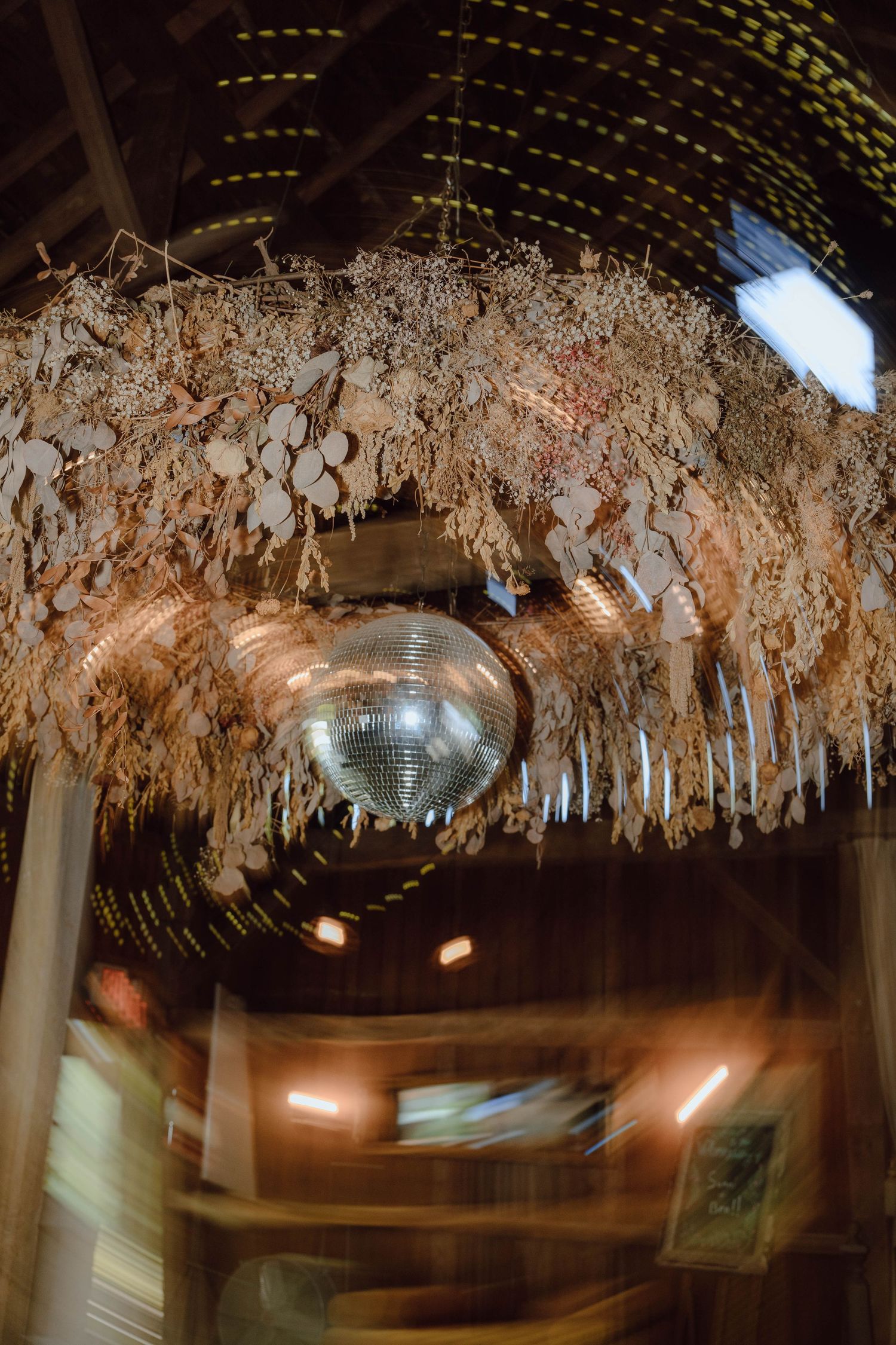 Disco ball hanging from a rustic barn ceiling adorned with dried florals and warm string lights.
