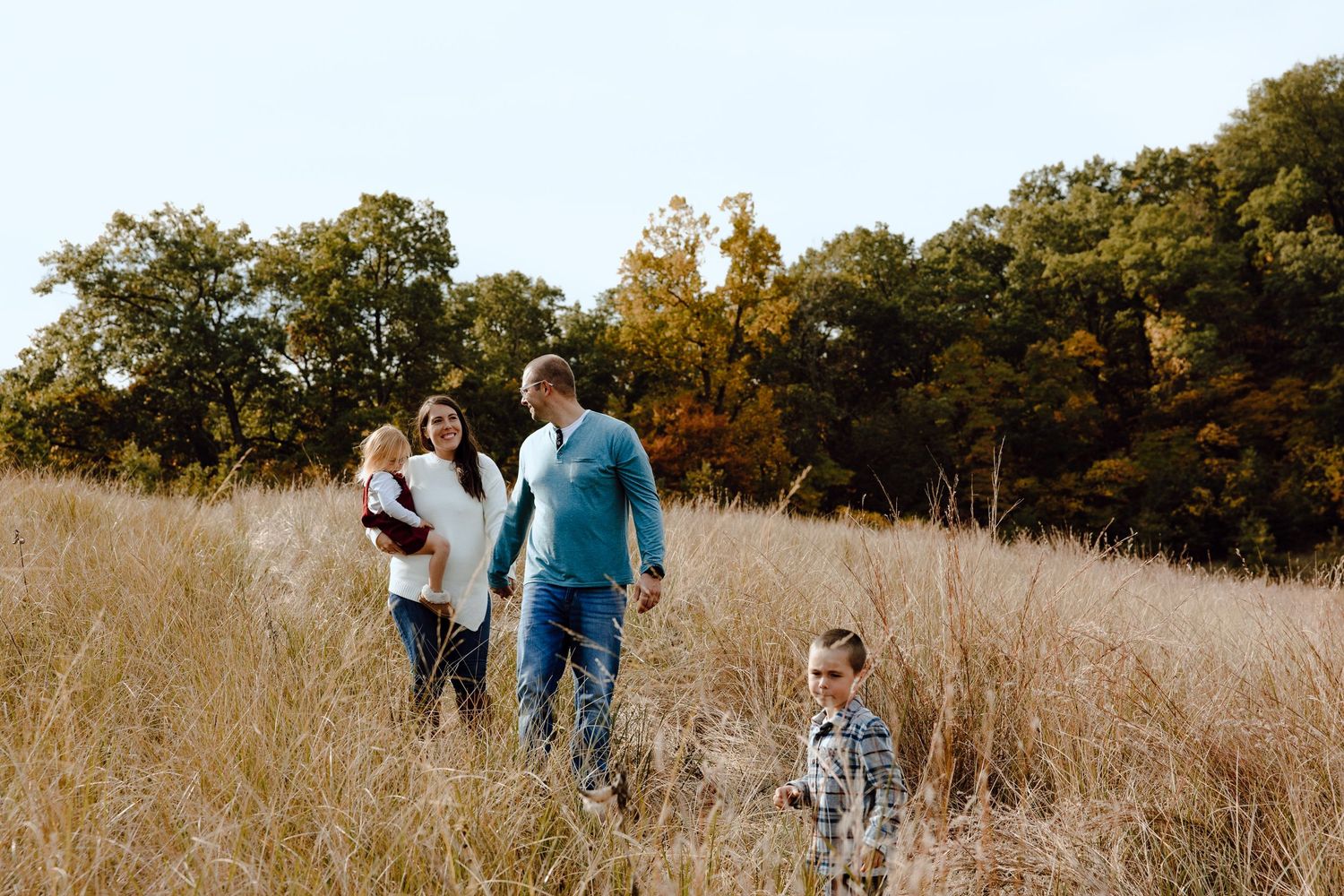 A family walks together through tall golden grass with autumn trees in the background.