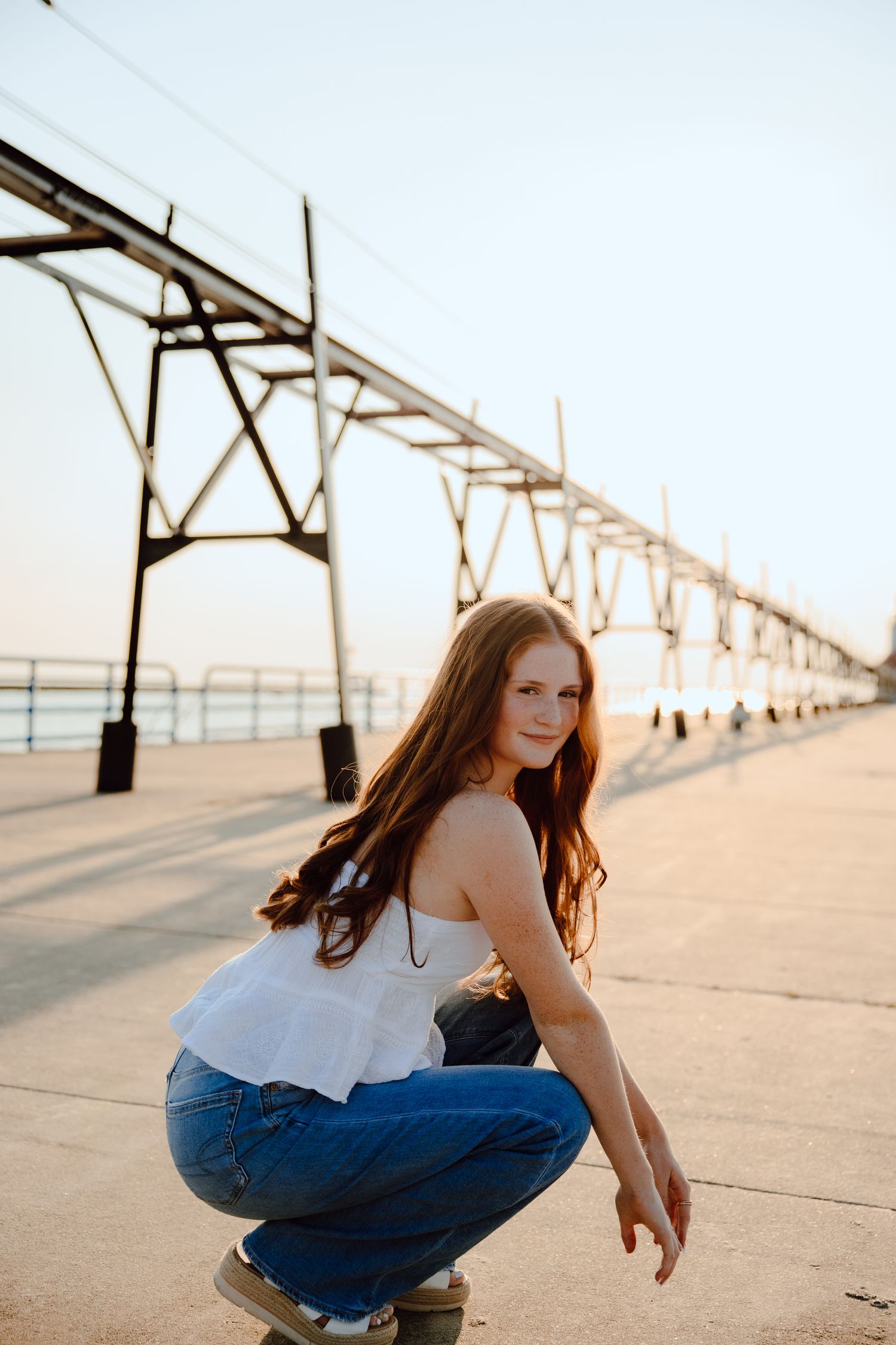 Someone in jeans and white top crouches on a pier during golden hour.