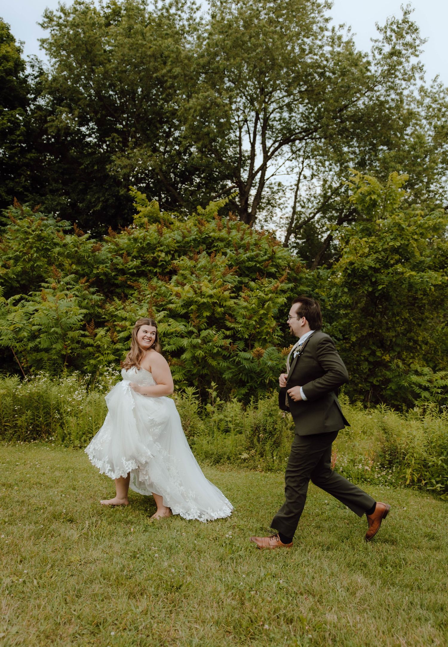 Bride and groom playfully running across green lawn in lush garden setting on wedding day.
