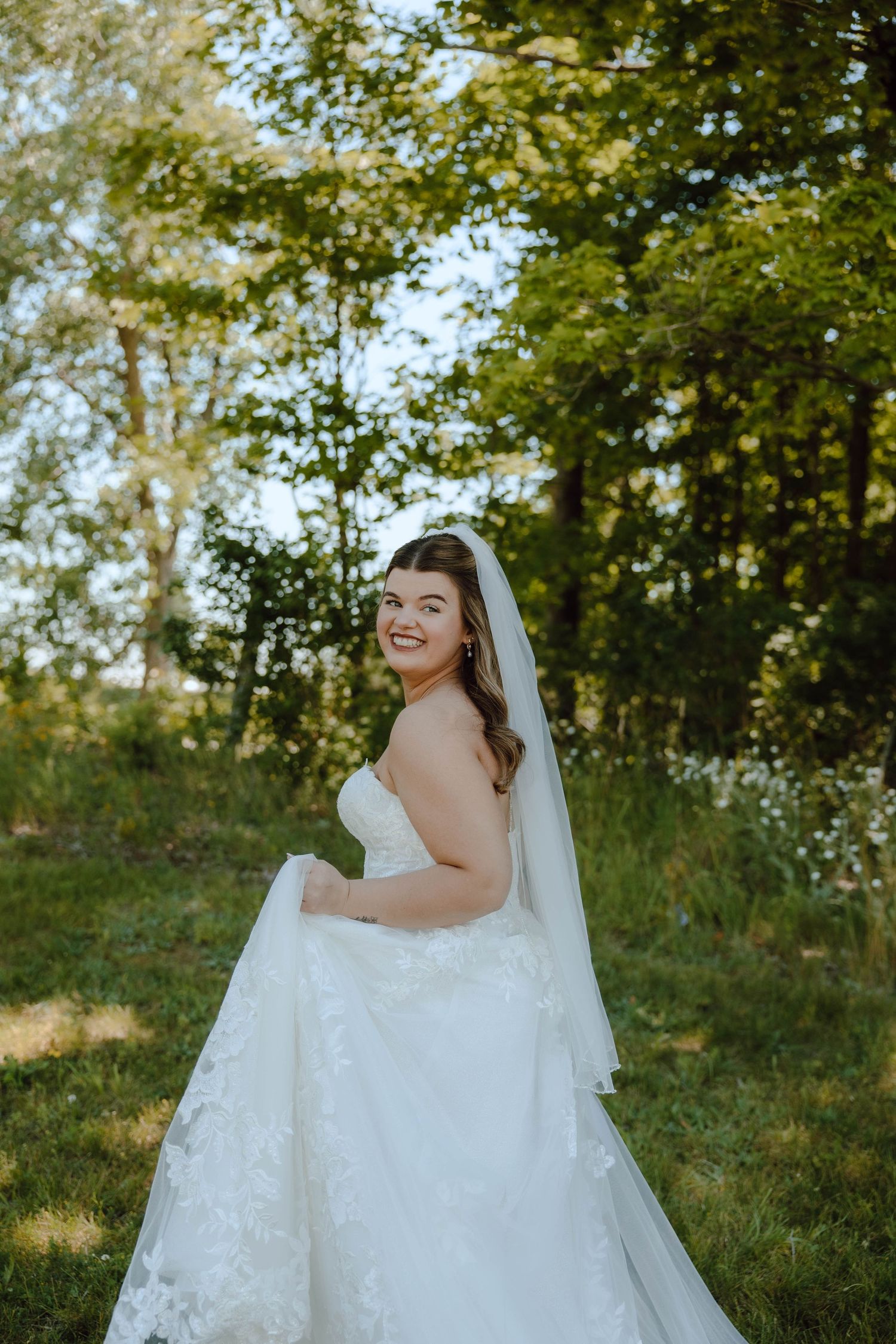 Smiling bride in strapless lace wedding gown with veil poses outdoors in sunlit green garden.