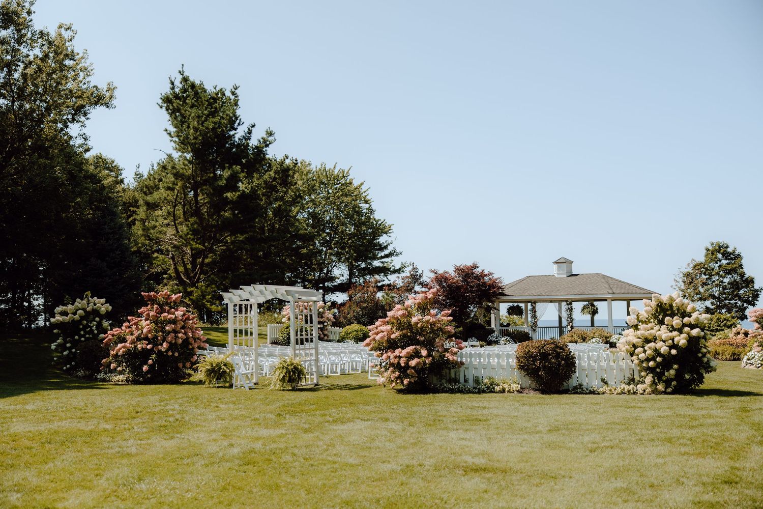 Elegant outdoor wedding venue with white gazebo, lush gardens, and blooming shrubs on a sunny day.