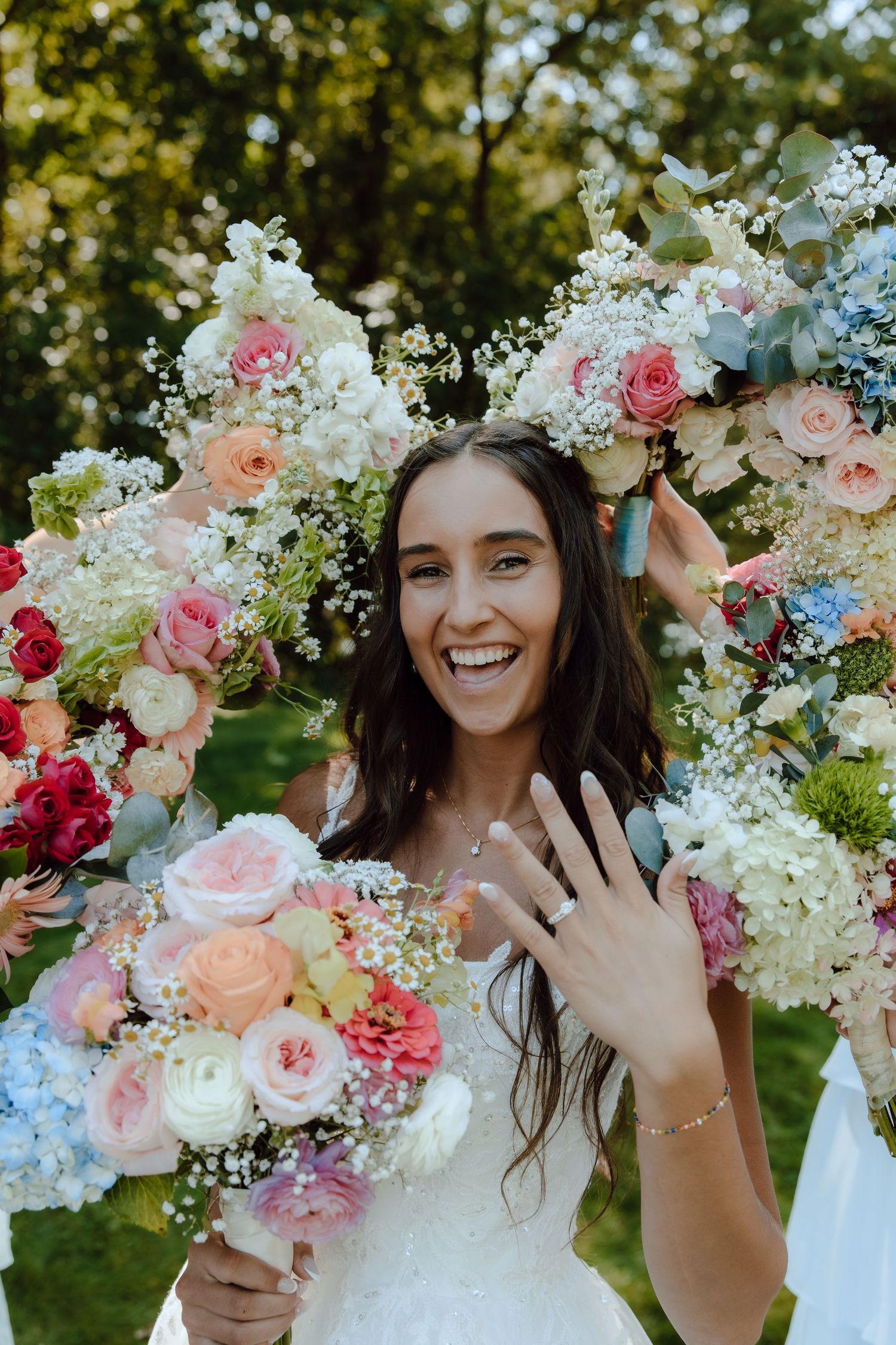Smiling bride showing wedding ring surrounded by colorful floral bouquets outdoors.