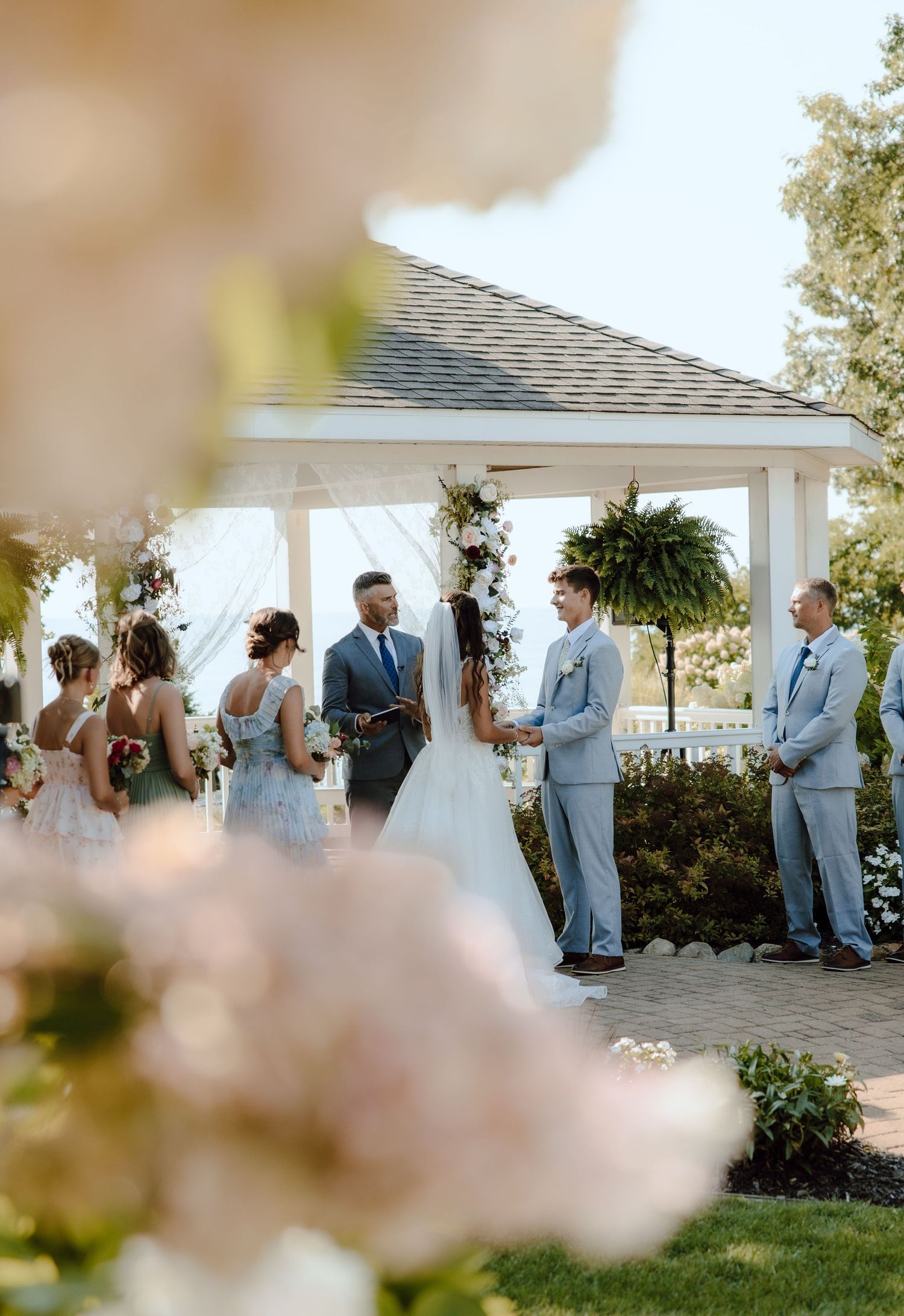 Wedding ceremony at outdoor gazebo with bridal party exchanging vows surrounded by flowers.