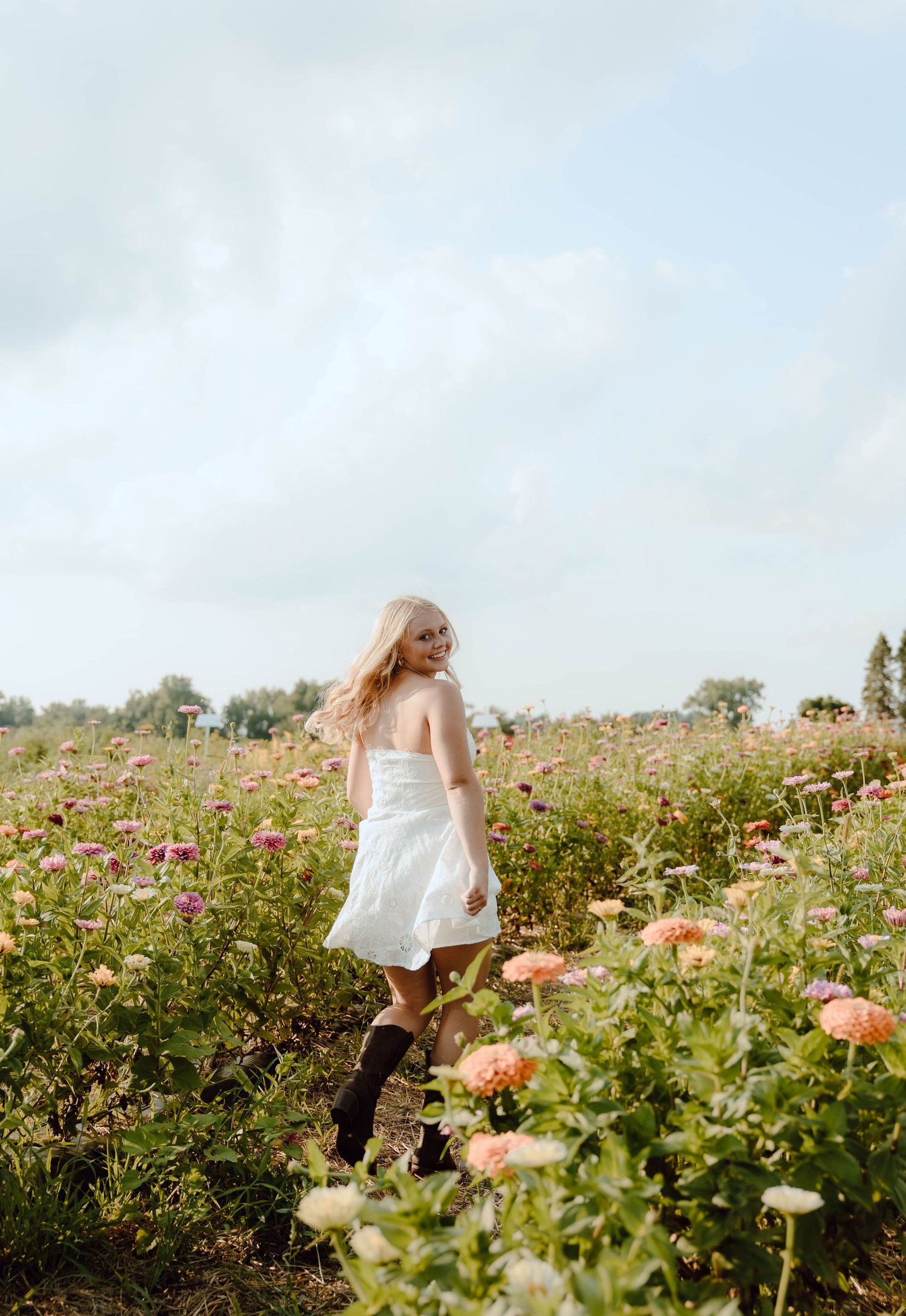 A blonde woman in a white dress and black boots walks through a vibrant flower field under a cloudy sky.