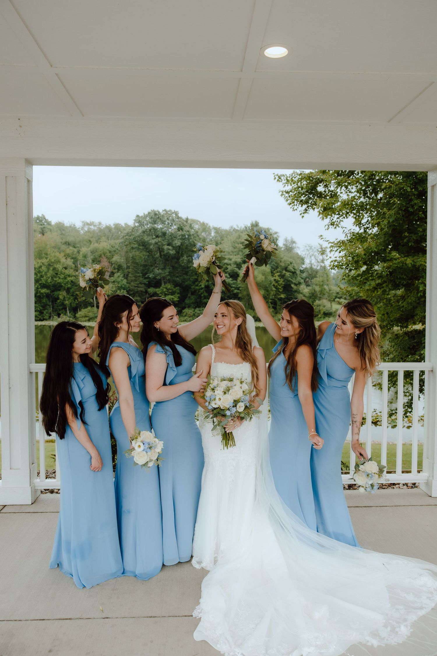 Bride in white gown holds bouquet surrounded by bridesmaids in blue dresses raising flowers on a porch.