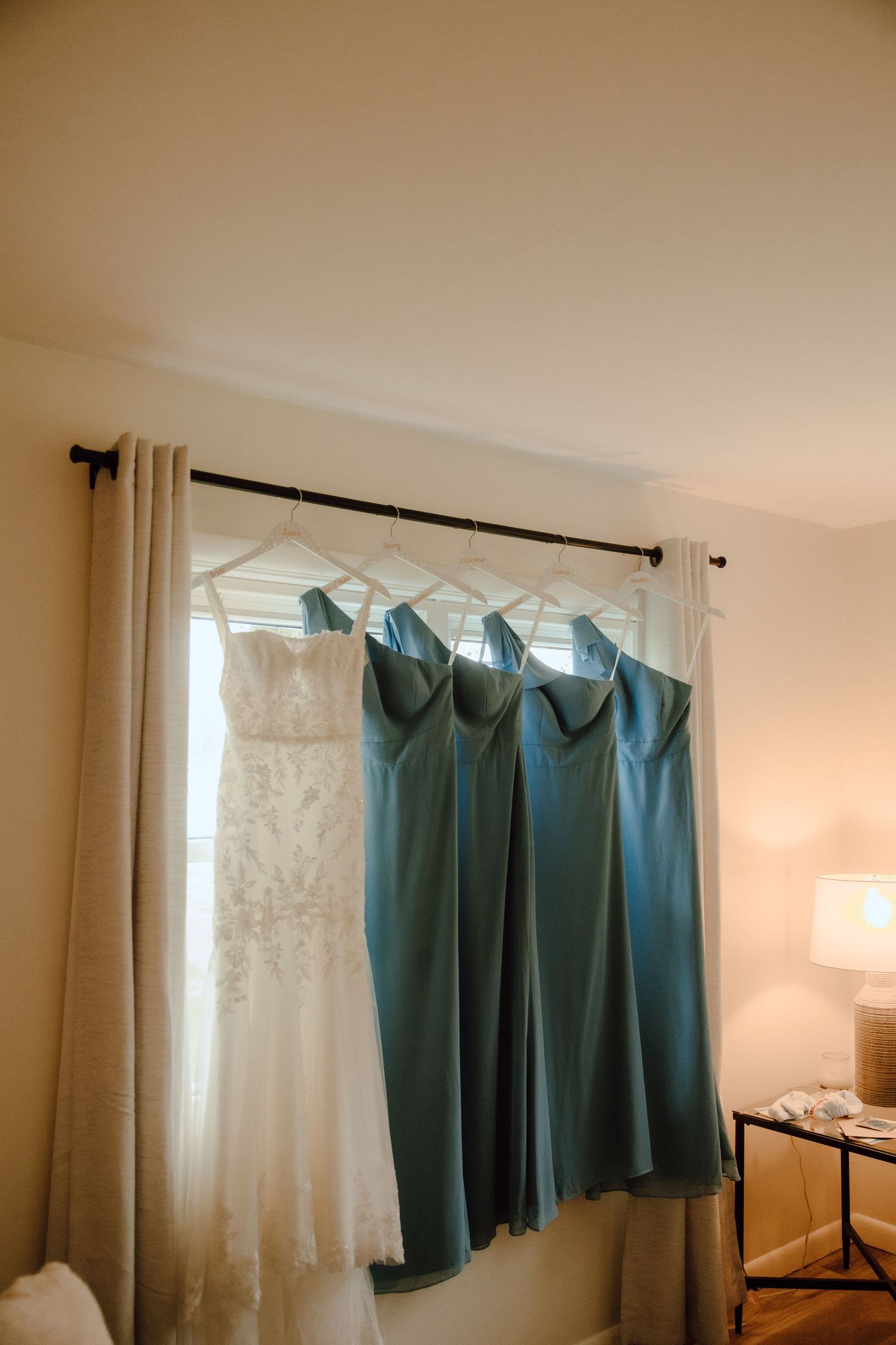 White wedding gown and teal bridesmaid dresses hanging on curtain rod by window.