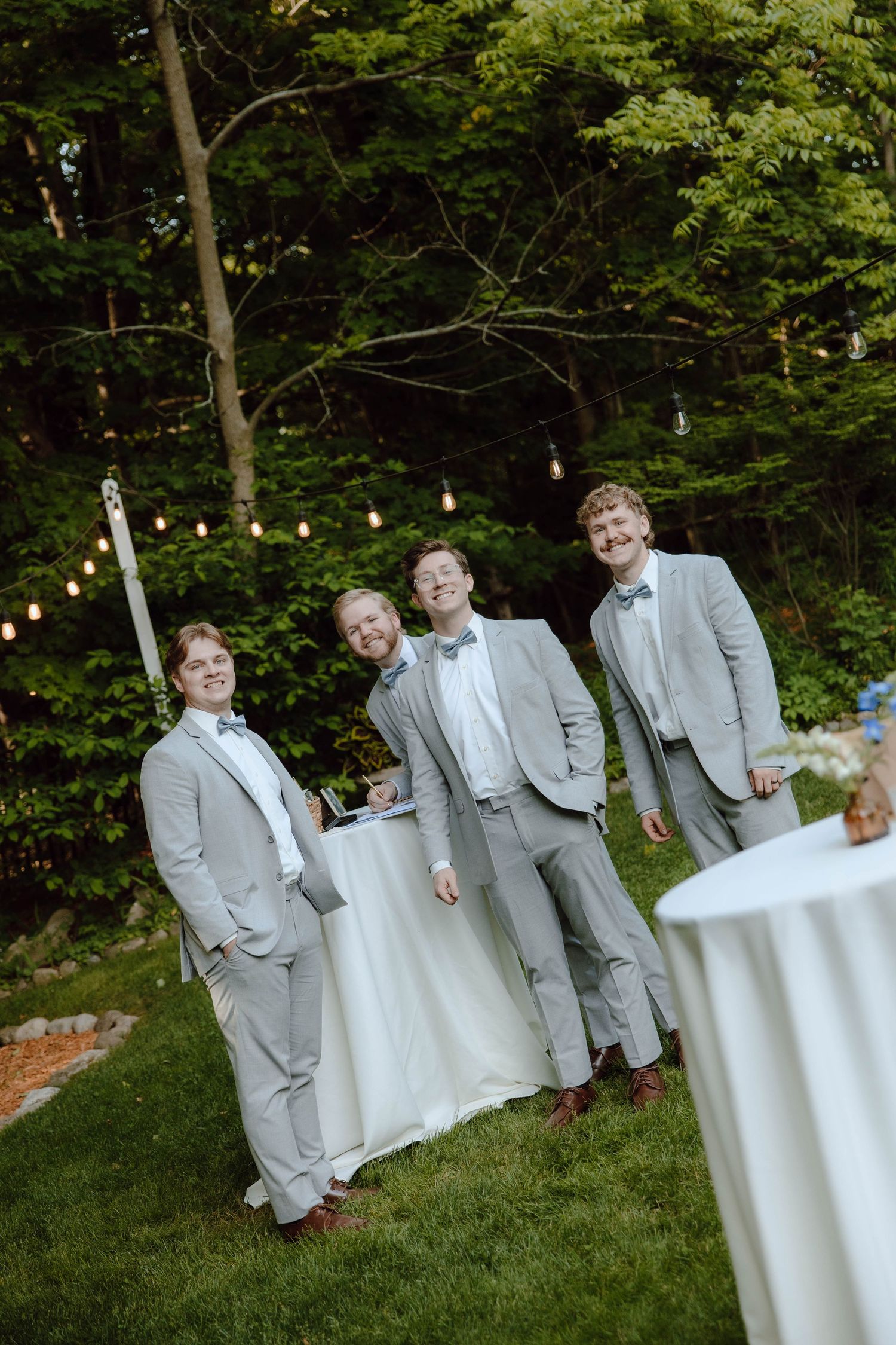 Three groomsmen in light gray suits with bow ties pose outdoors at a wedding reception with string lights.