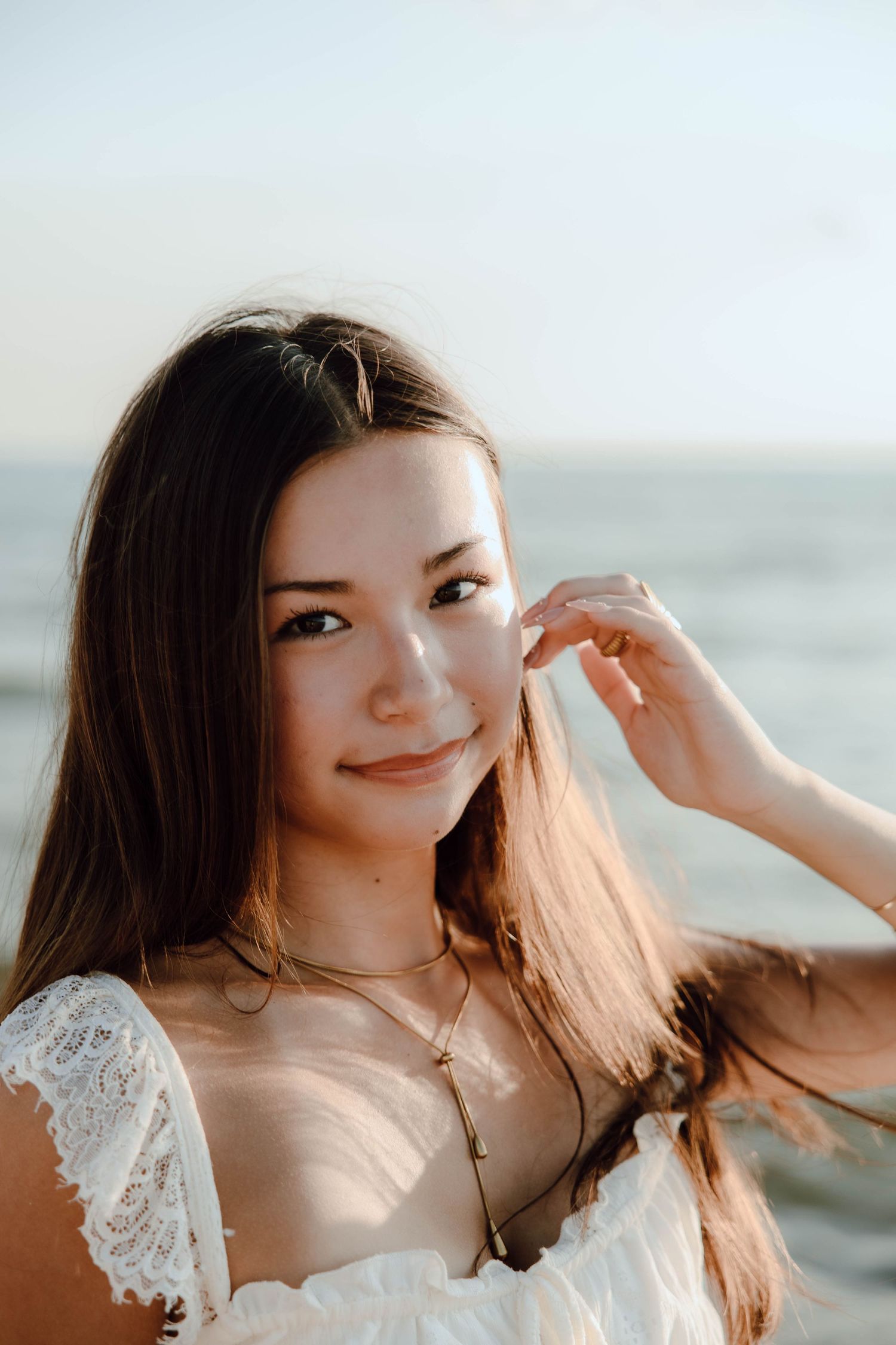 Young woman with long brown hair in white lace dress smiling at camera with ocean in background.