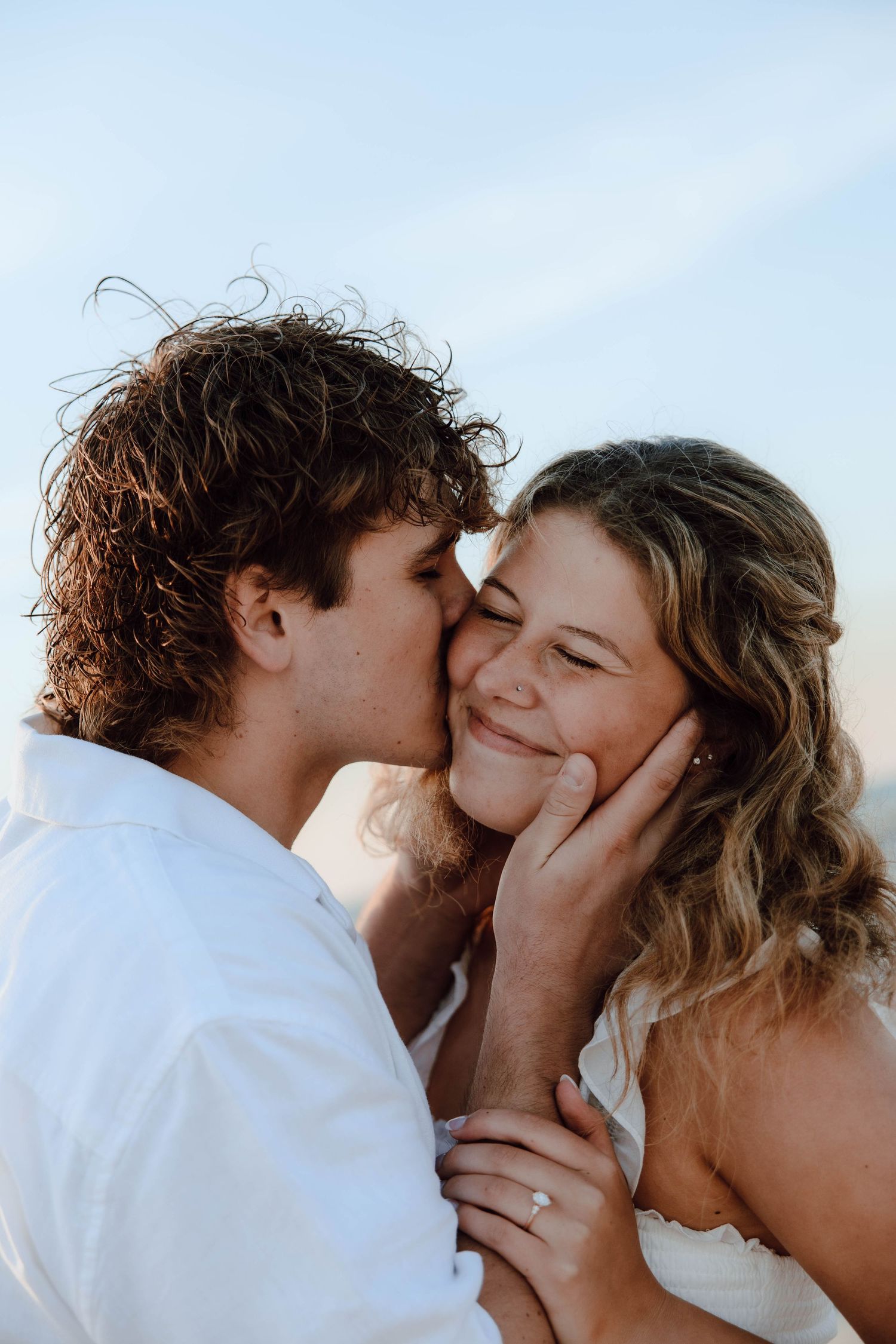 A couple shares a tender kiss on the cheek outdoors, the woman smiling and wearing an engagement ring.