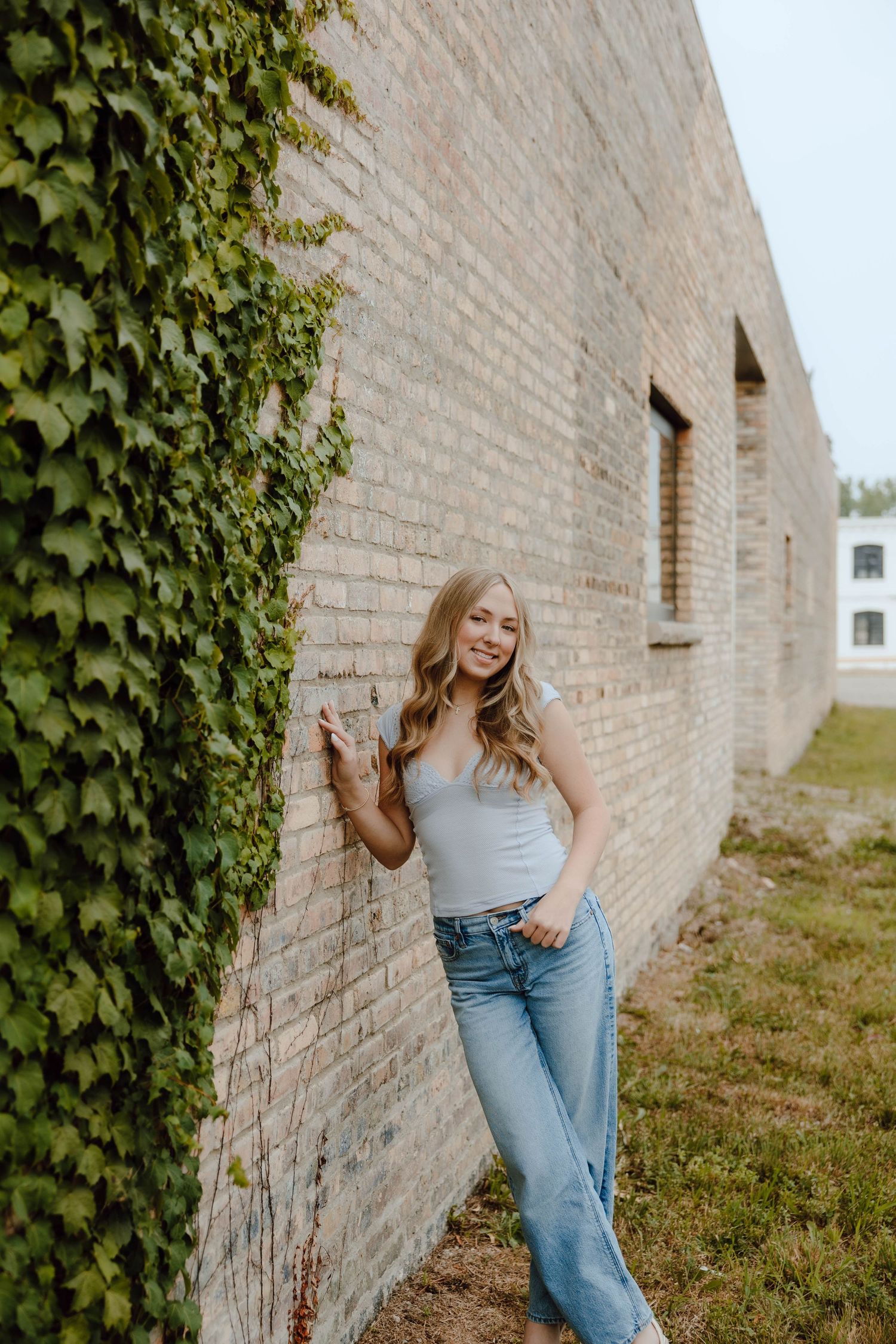 Young woman in blue jeans and gray tank top leaning against brick wall with ivy, smiling outdoors.