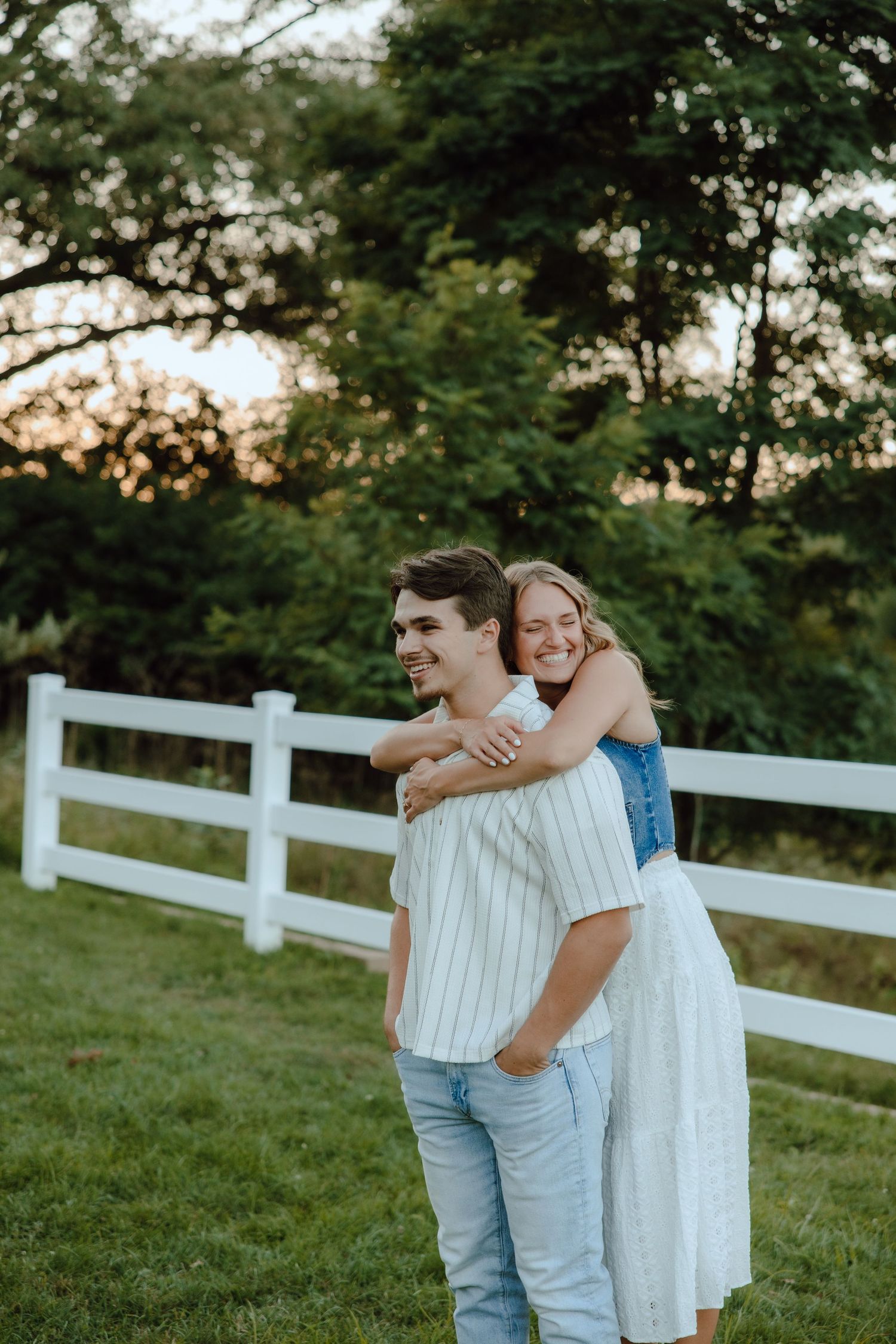 A smiling couple embracing outdoors near a white fence at sunset, woman hugging man from behind.