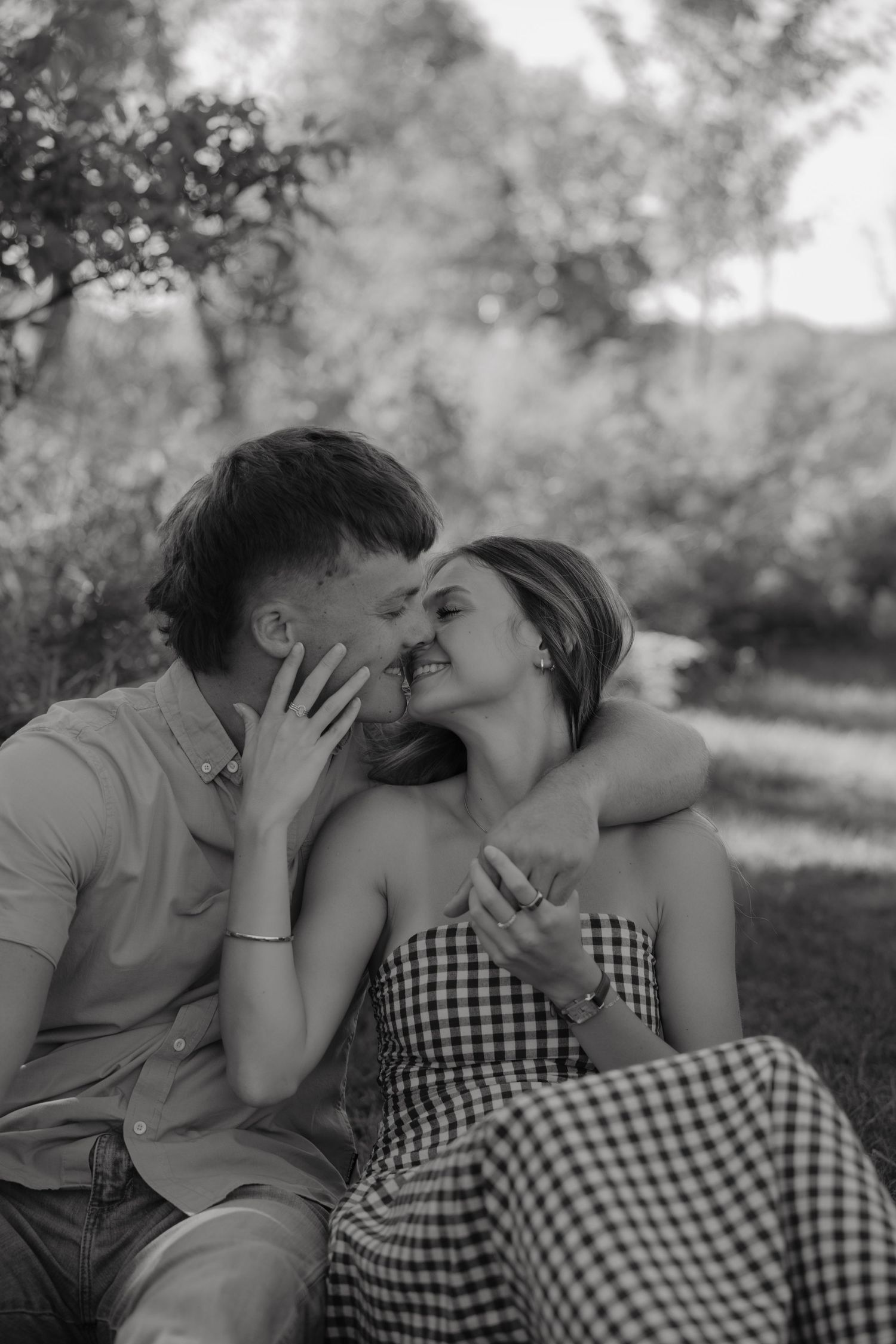 Black and white photo of a romantic couple sitting outdoors, sharing a tender moment as they lean in for a kiss.