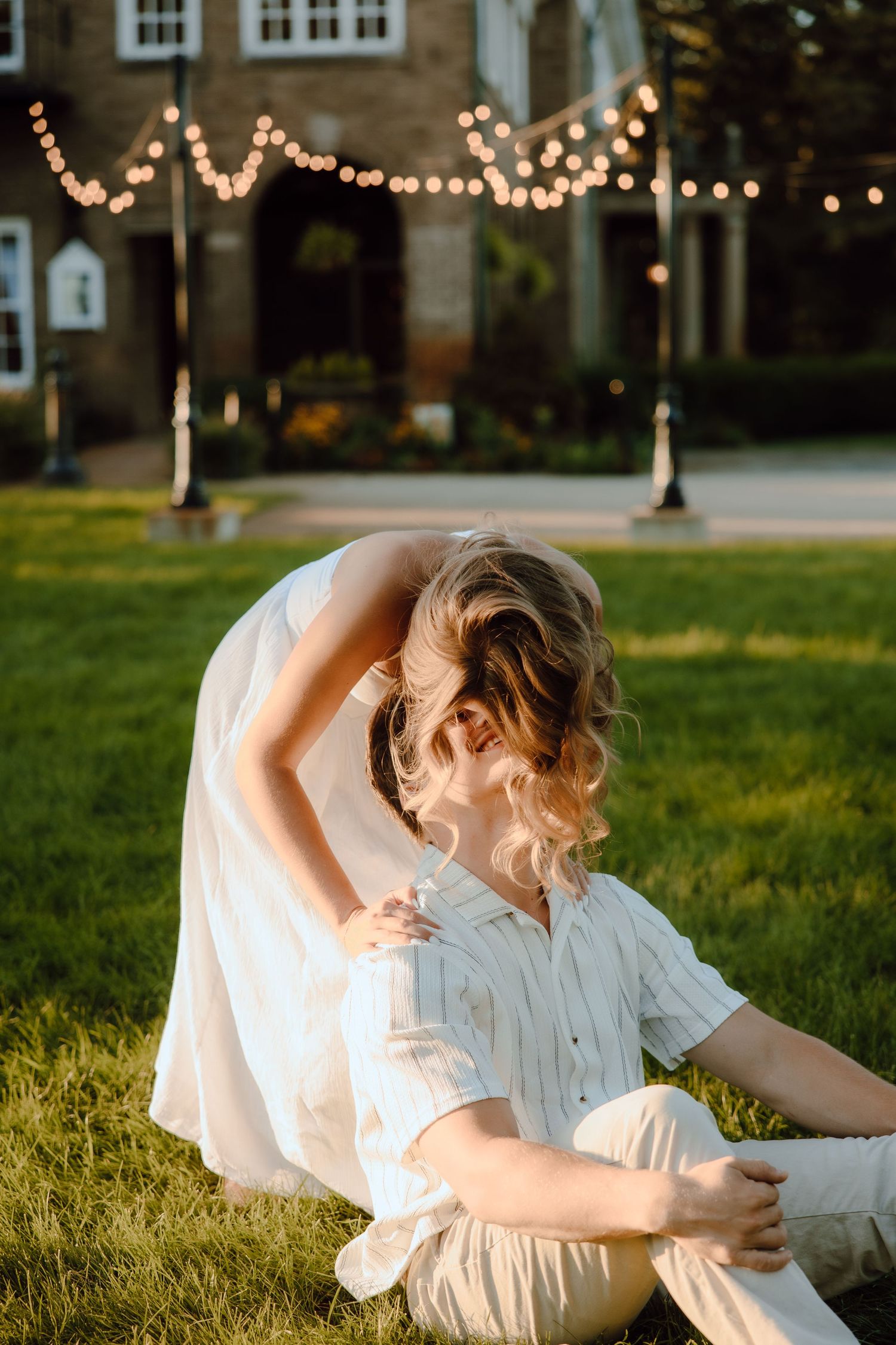 Couple in white outfits sitting on grass at golden hour, with string lights and brick building backdrop.