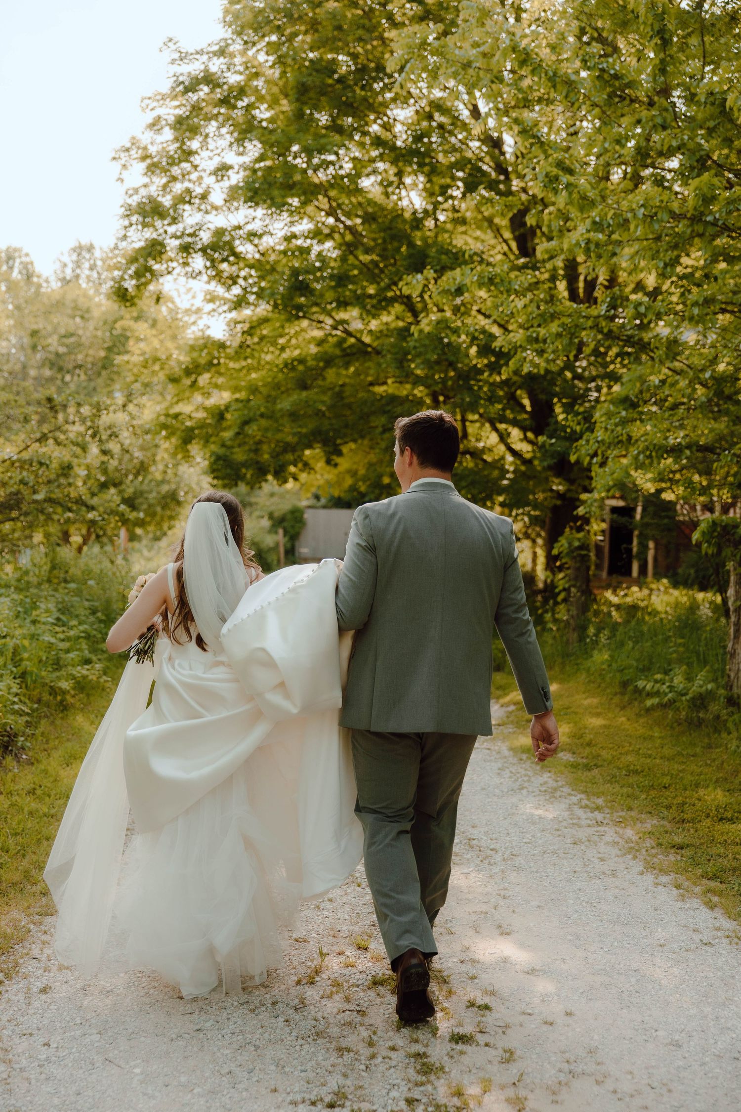 Bride and groom walking hand in hand down a tree-lined gravel path on their wedding day.