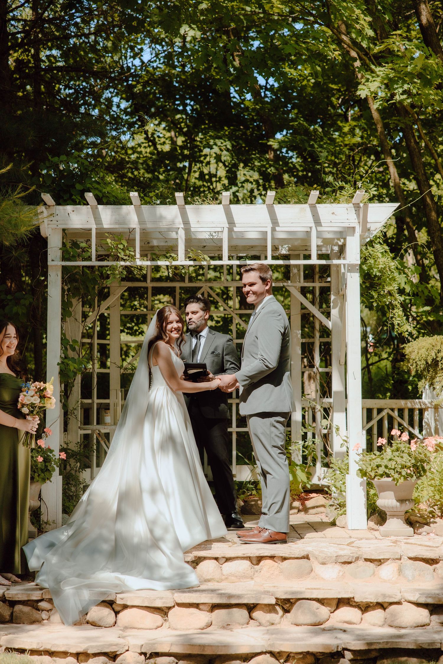 Bride and groom exchange vows outdoors under a white pergola arbor surrounded by lush green trees on their wedding day.