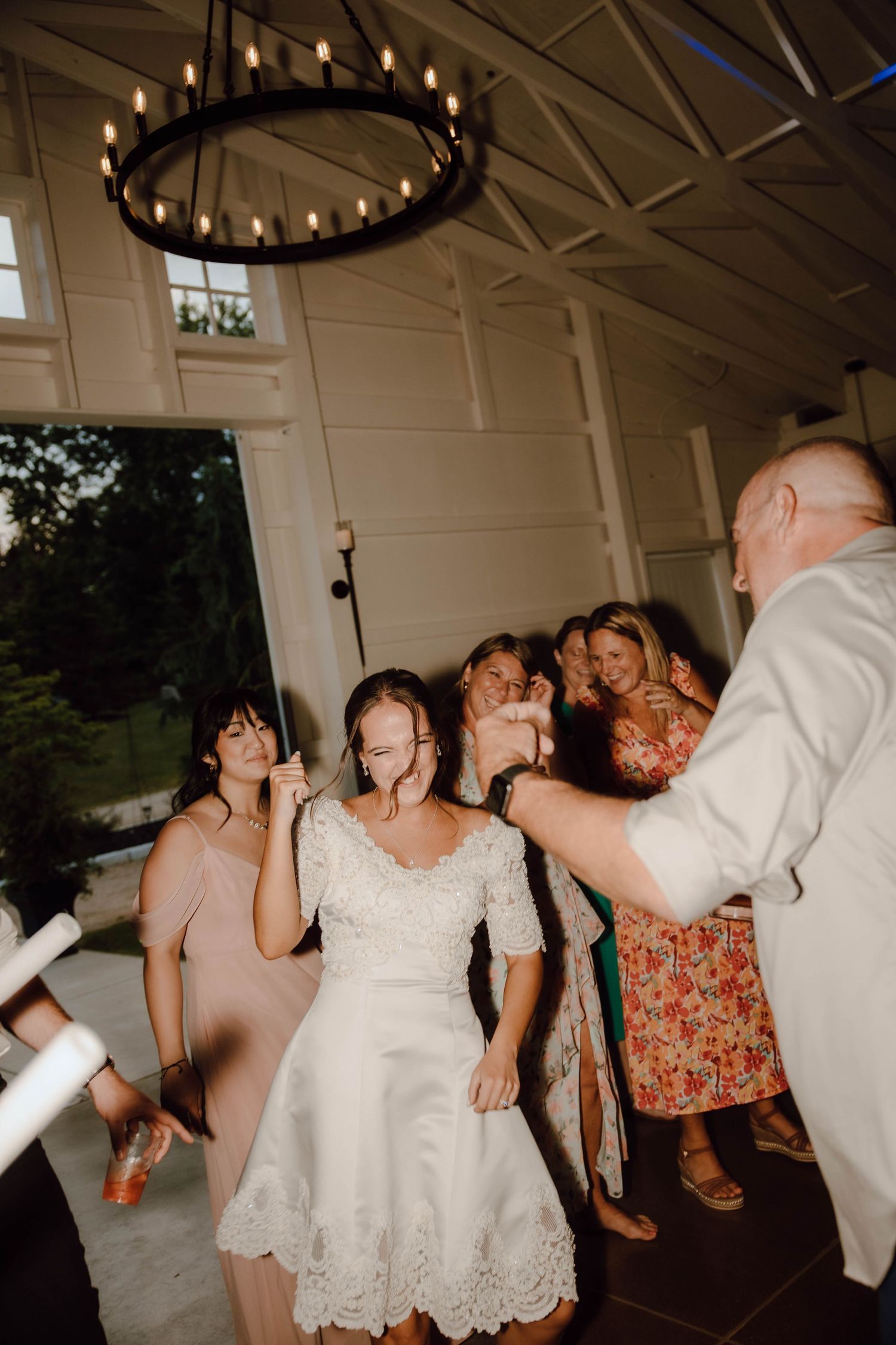 Bride in lace dress dancing joyfully with wedding guests under chandelier at elegant indoor reception.