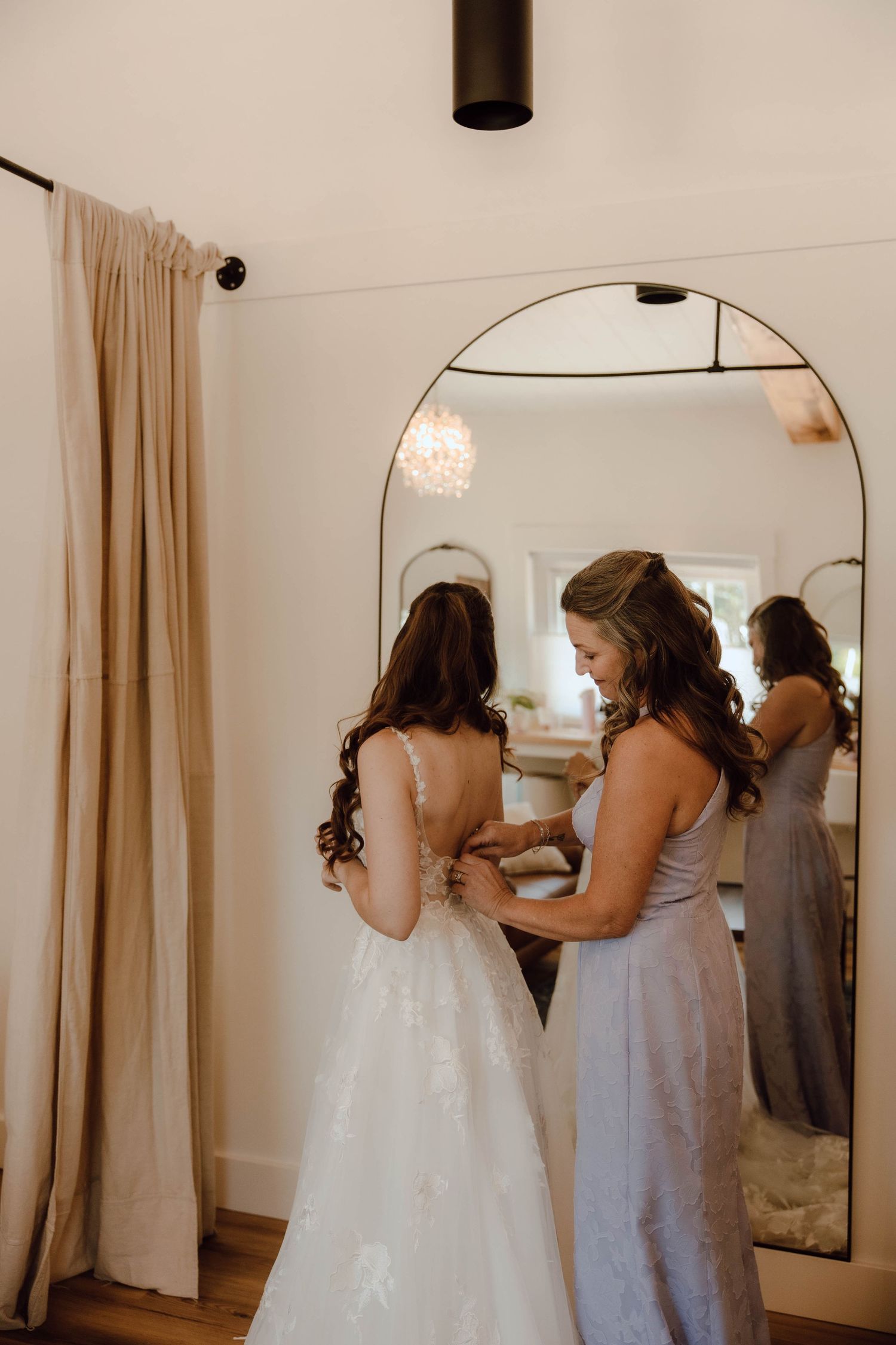 Bridesmaid helping bride fasten her lace wedding gown in a bright dressing room with an arched mirror reflection.