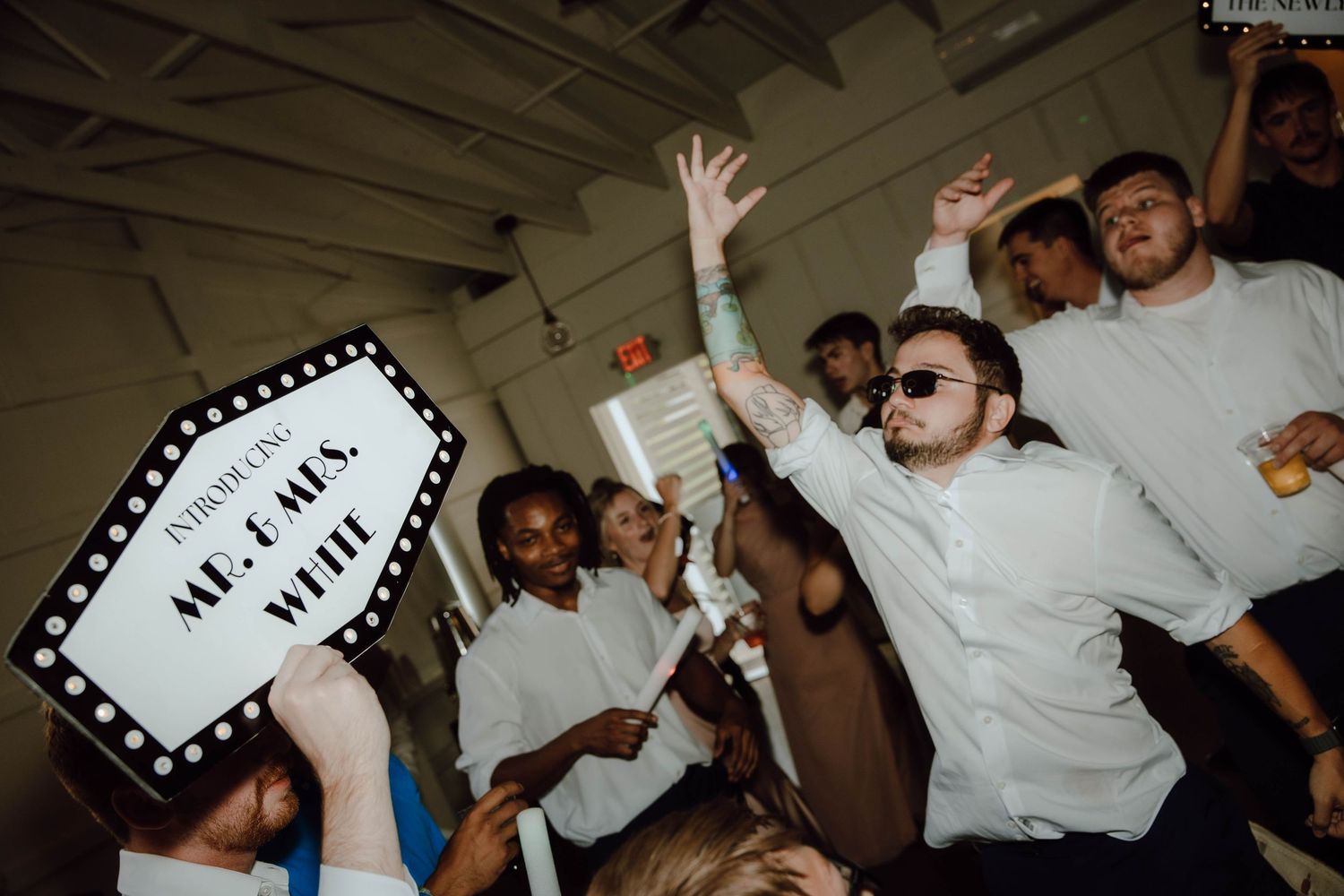 Guests dancing at a lively wedding reception, man in white shirt raising arm, holding Mr. & Mrs. sign.