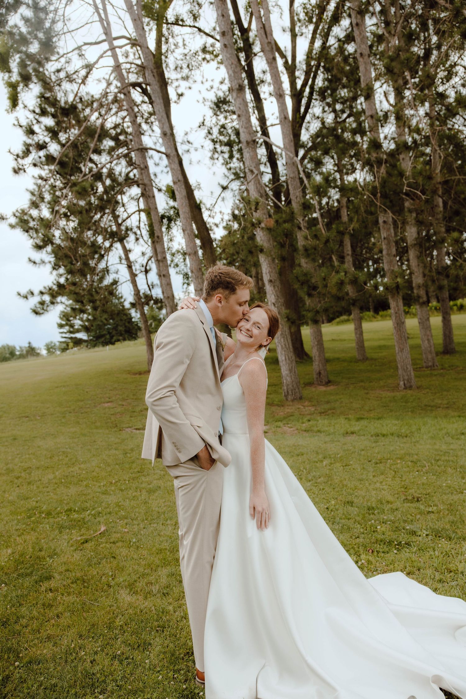 Groom in tan suit kissing bride in white gown outdoors on lush green lawn surrounded by tall trees.