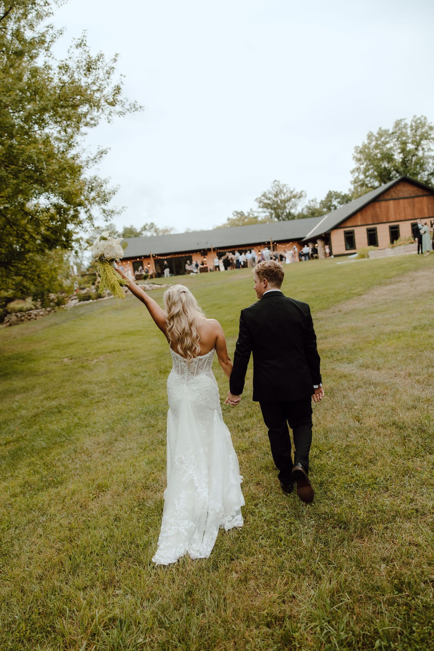 Bride and groom walk hand in hand toward barn venue, bride raising bouquet in celebration.
