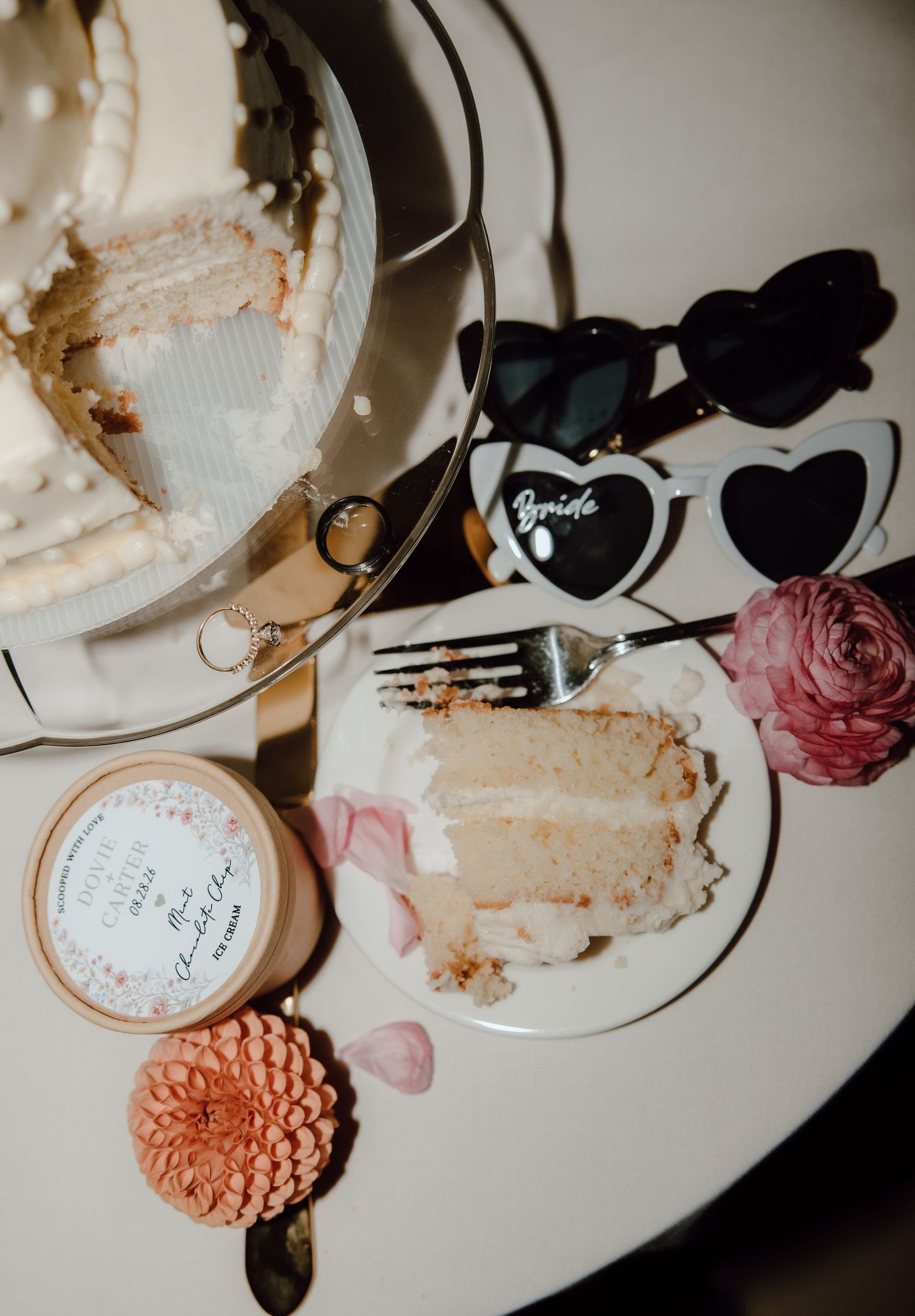 Elegant wedding cake slice on a white plate beside heart-shaped bride sunglasses, pink flowers, and a personalized favor box.