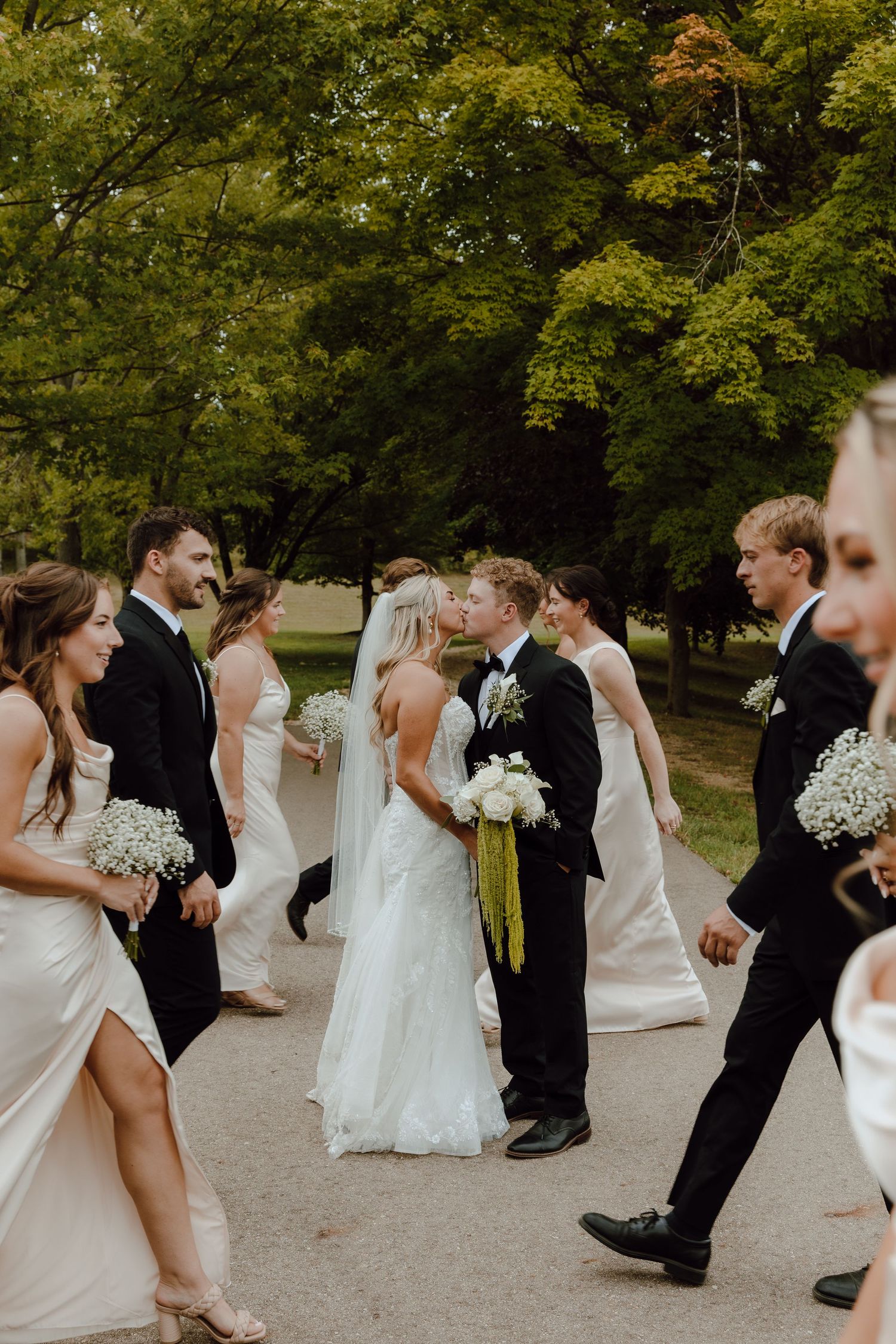Bride and groom share a kiss surrounded by wedding party in white gowns and black tuxedos on a tree-lined path.