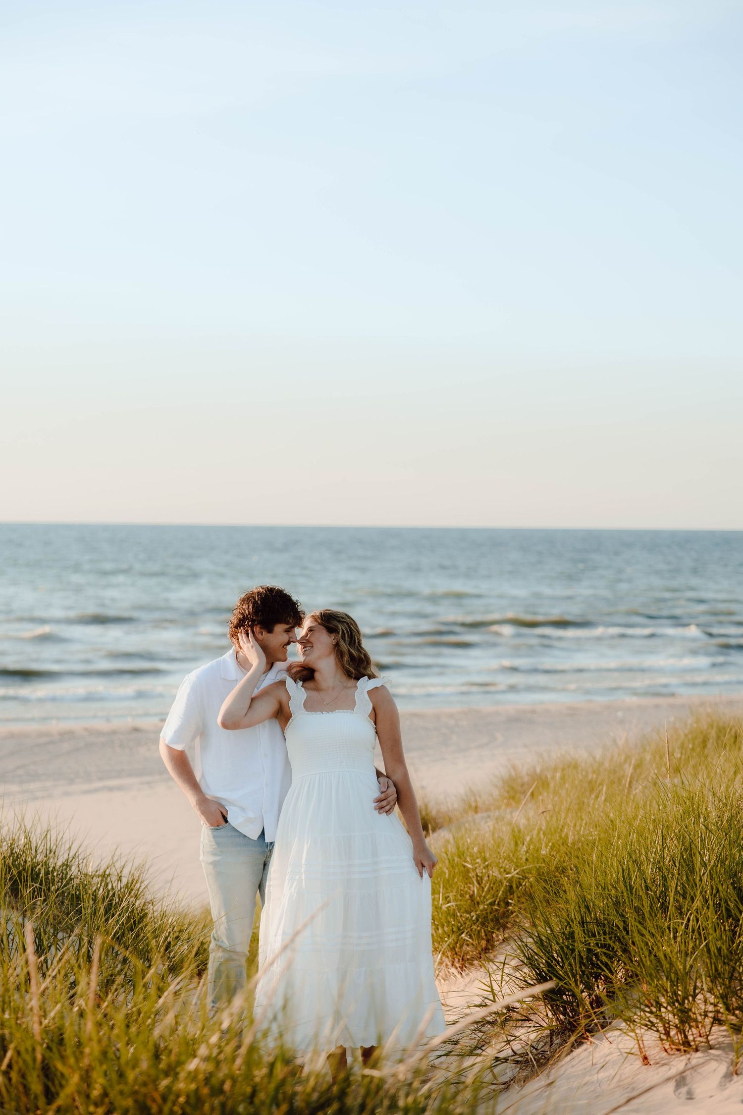 Couple sharing a romantic moment on a sandy beach with ocean waves and grass dunes in warm golden light.