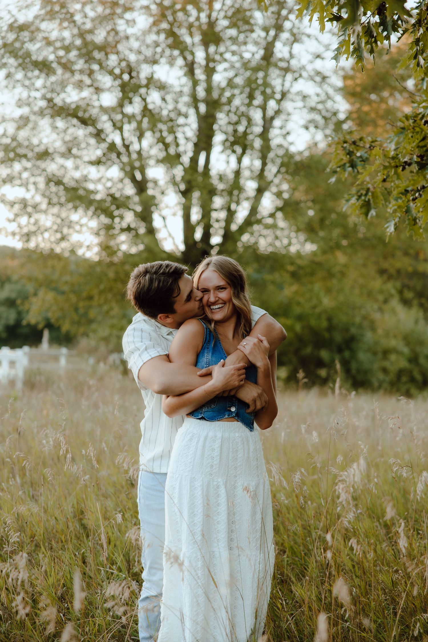Man kissing smiling woman on the cheek while embracing in a tall grass field with trees behind.