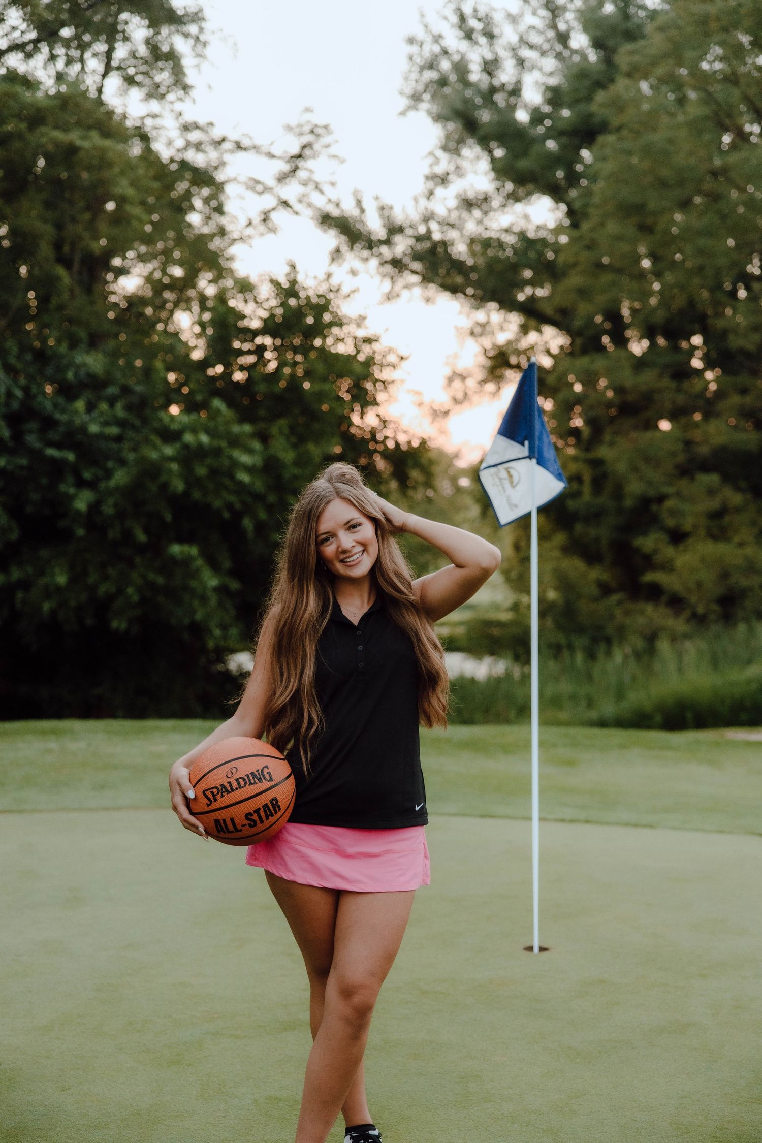 Young woman in pink skirt holding a Spalding basketball on a golf course green with flag.