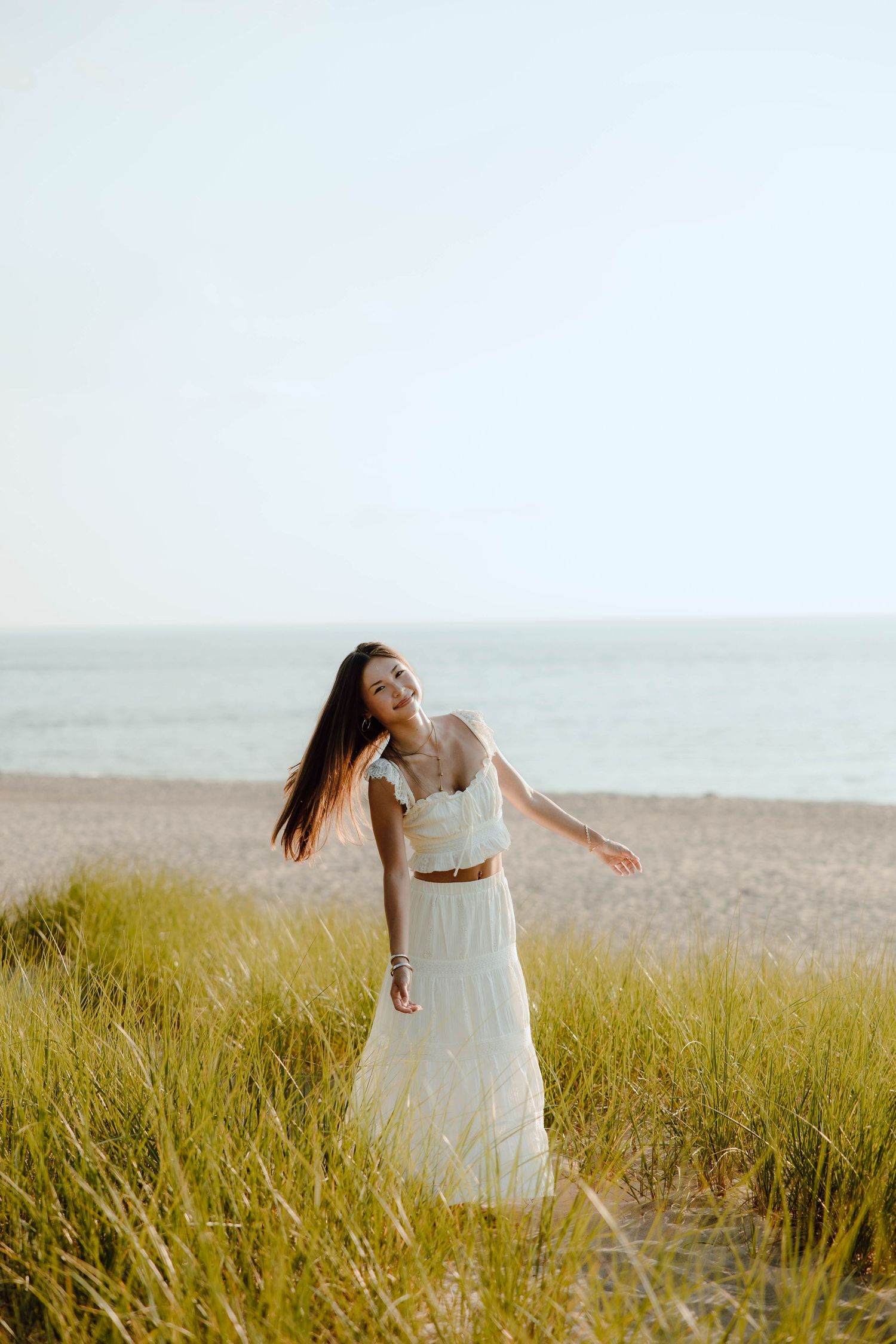 Woman in white crop top and maxi skirt posing gracefully among beach grass with ocean in background.