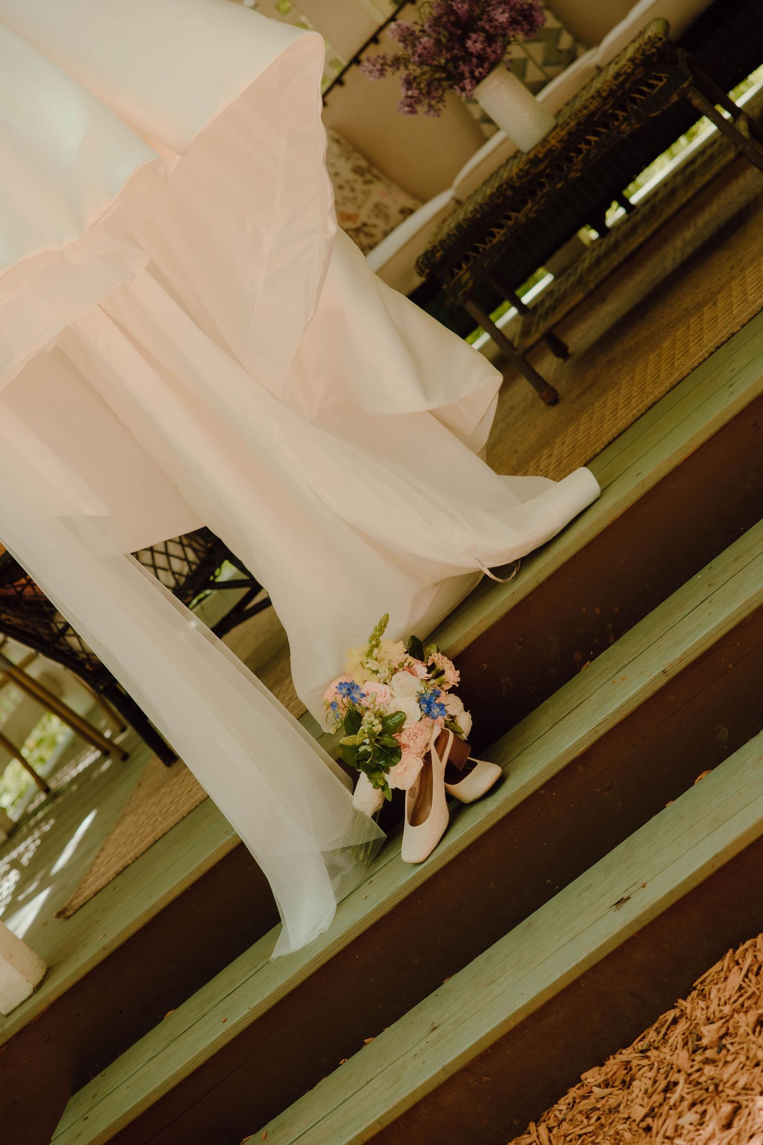 White bridal shoes and floral bouquet on wooden steps beside flowing pink fabric at a wedding venue.