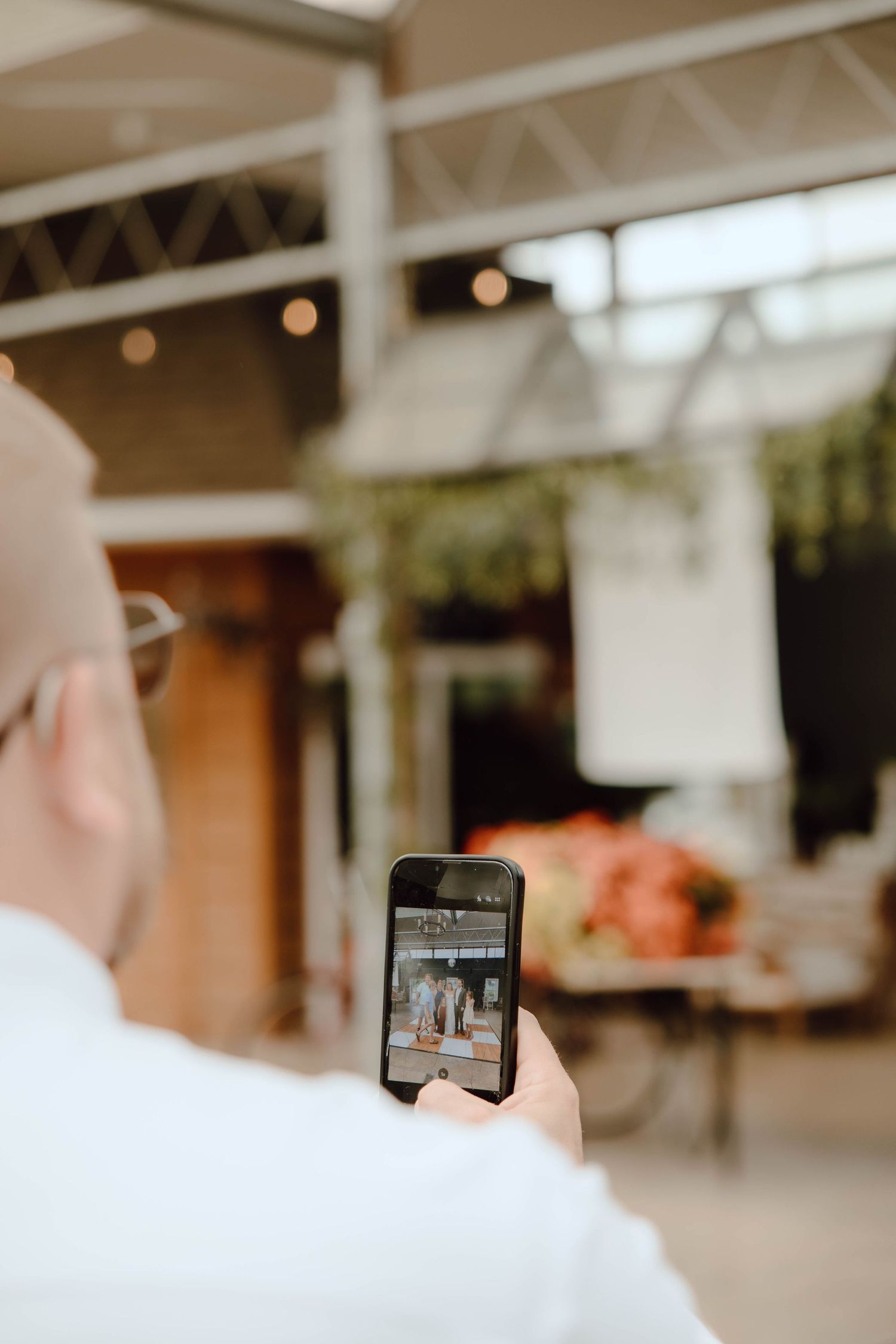 Man holding smartphone photographing a decorated event venue with floral arrangements and string lights.