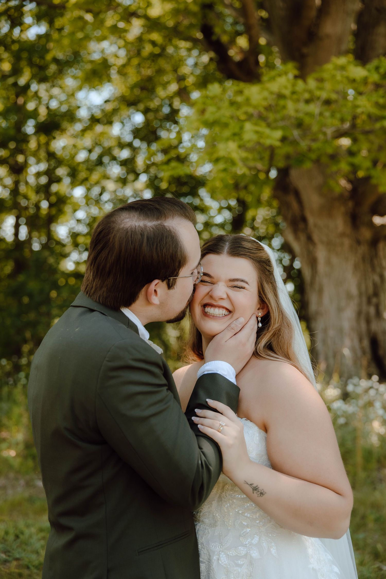 Groom in olive suit kisses laughing bride on cheek outdoors near a large tree.