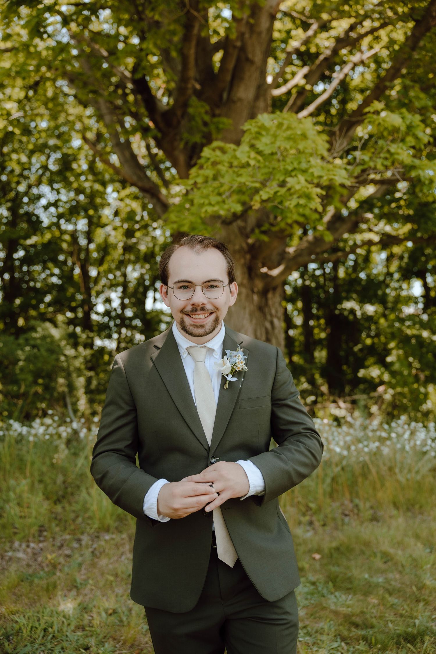 Smiling groom in olive green suit with boutonniere stands outdoors before a large tree on his wedding day.