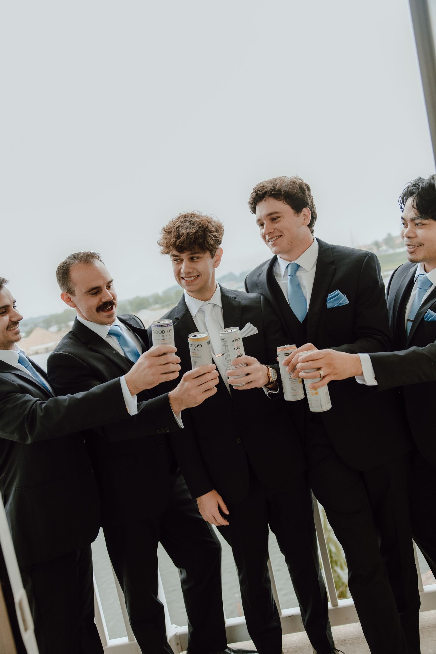 Groomsmen in black suits with light blue ties toasting with canned drinks on a balcony before a wedding ceremony.