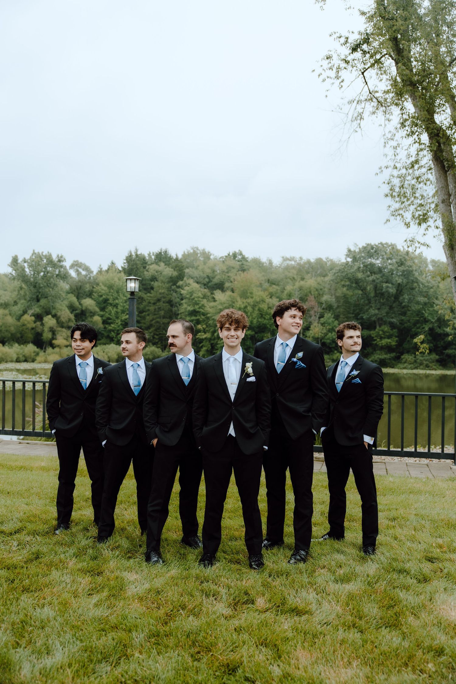 Groom and six groomsmen in black suits with blue ties pose outdoors near a pond and lush green trees.