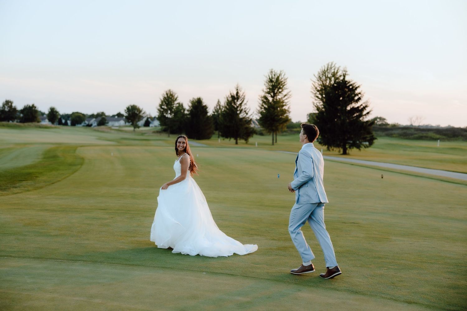 Bride in white gown and groom in light blue suit walk together on a golf course at sunset.