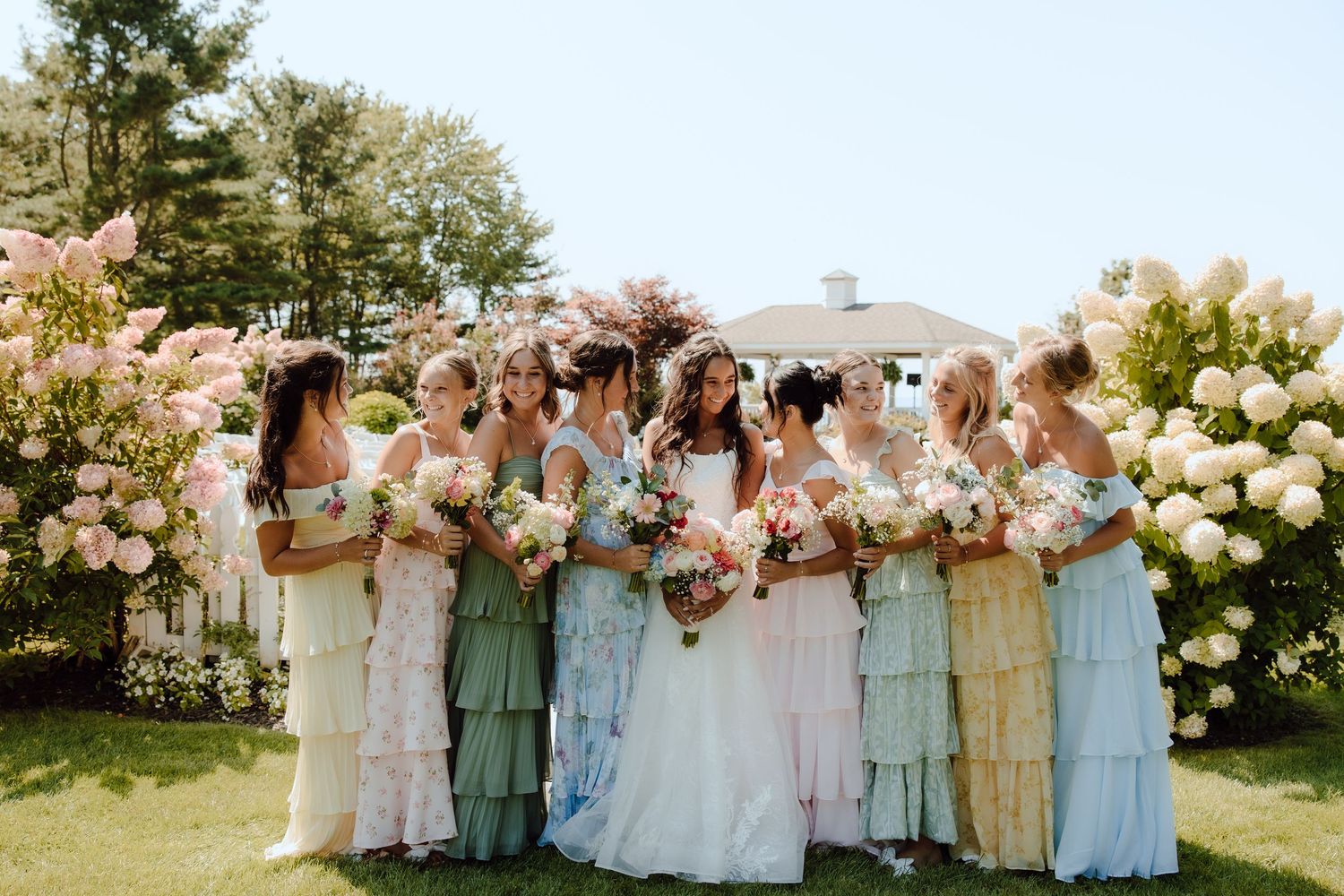 Bride surrounded by bridesmaids in mismatched pastel dresses near blooming hydrangea bushes.