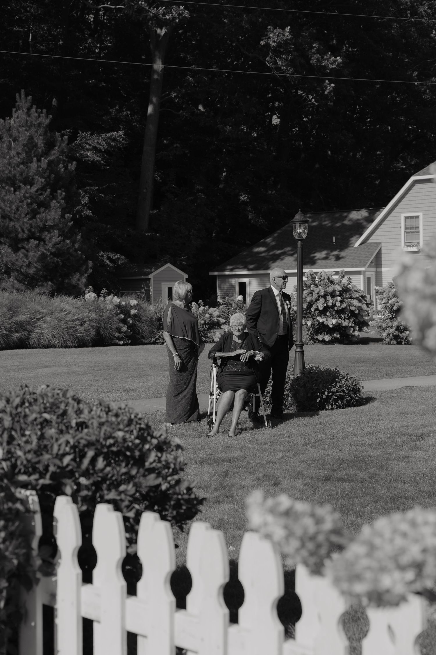 Black and white photo of wedding guests escorting elderly woman in wheelchair across a manicured lawn.
