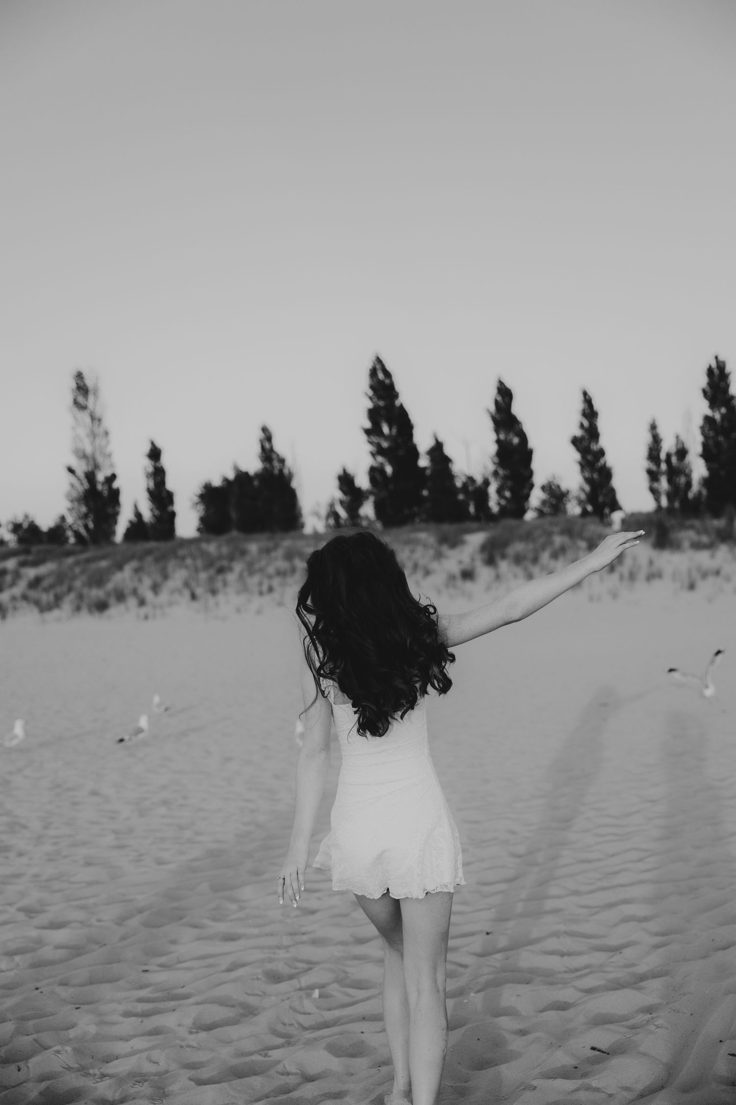 Black and white photo of someone twirling in a dress on sandy dunes with trees in background.