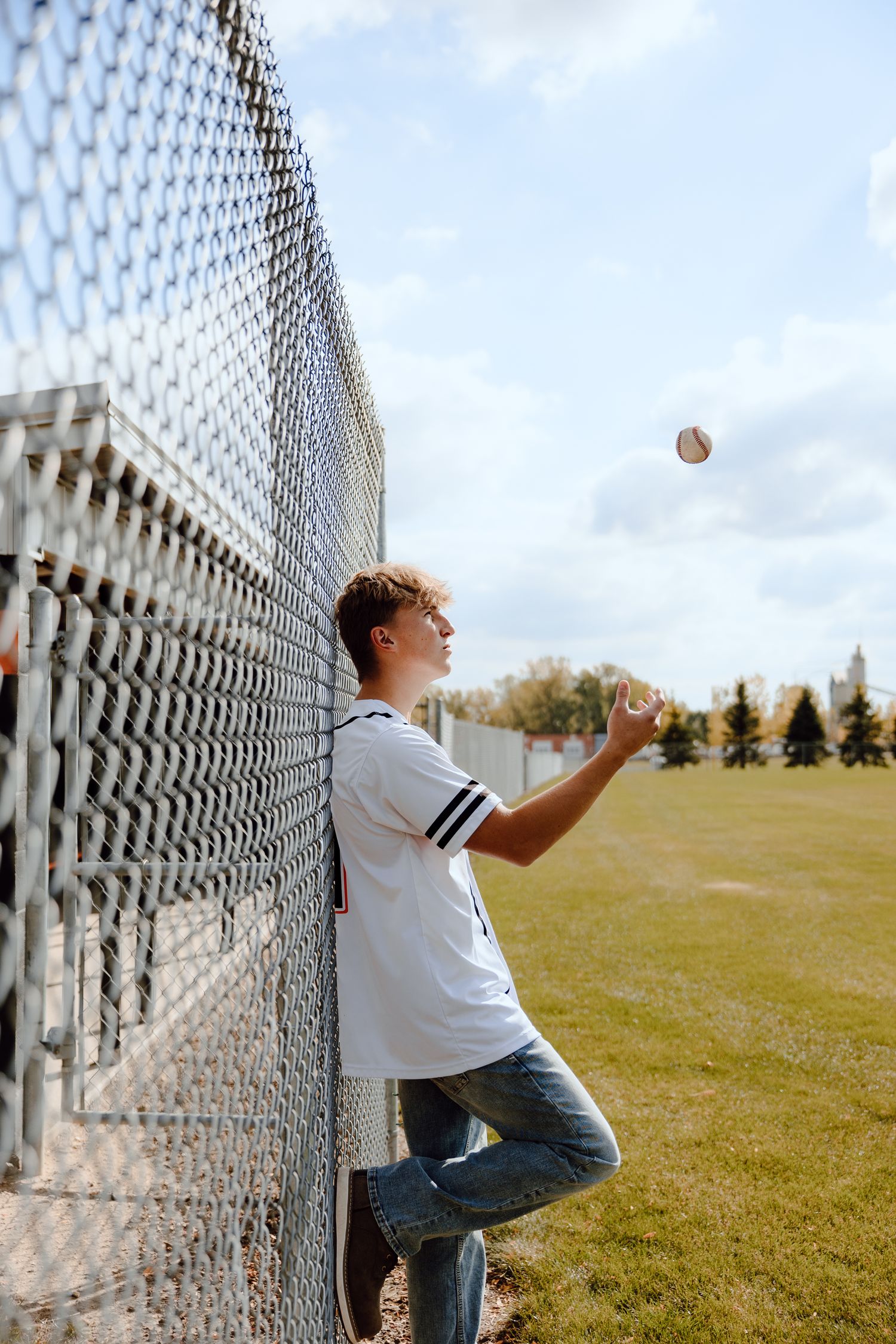 Baseball player leaning against chain link fence tossing ball in athletic field.